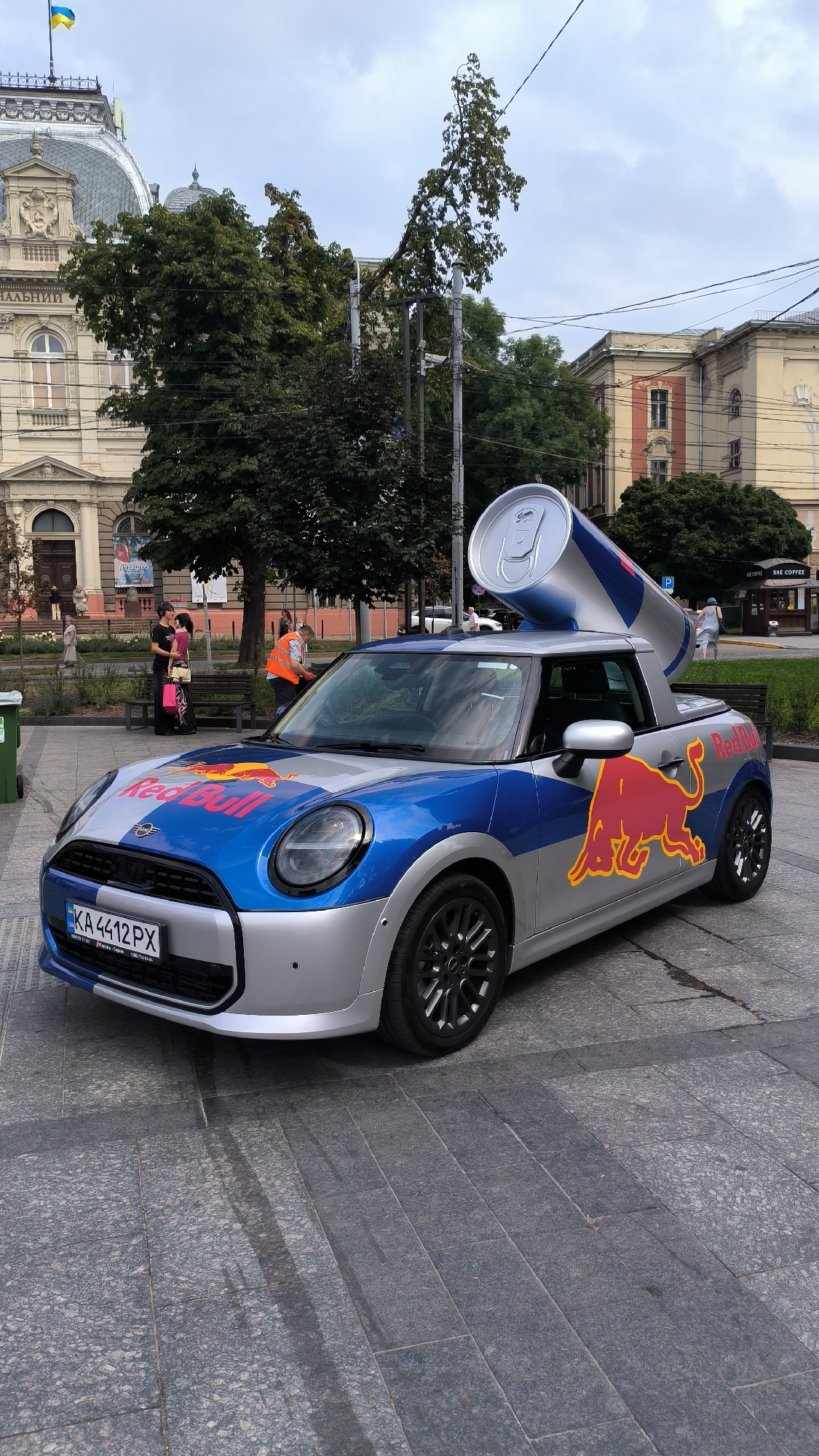 A Mini Cooper convertible, decorated with Red Bull branding and a giant Red Bull can on its roof, is parked on a stone-paved plaza in Lviv, Ukraine. The car is silver and blue, with the Red Bull logo prominently displayed on the hood and sides. The giant can, a replica of a Red Bull energy drink can, is tilted on the roof, with its opening facing forward.
In the background, a building with classical architecture and a dome, displaying a Ukrainian flag, is visible. Several people are walking around the plaza, and some are standing near benches. The weather appears overcast. The car's license plate reads "KA 4412 PX". The scene suggests a promotional event or a street display.