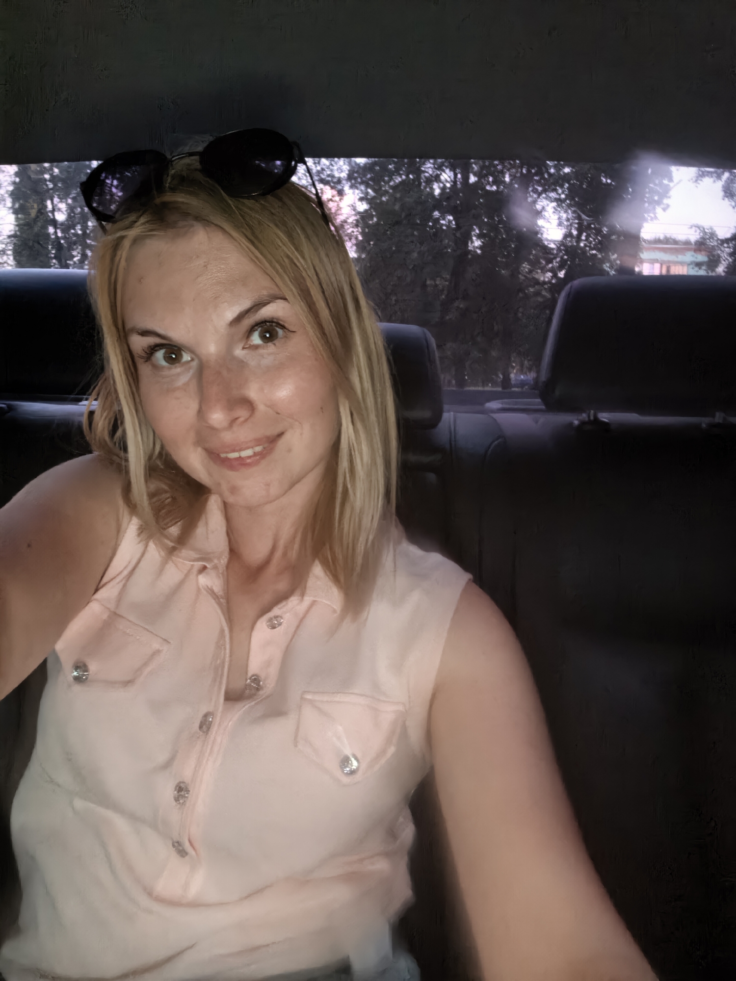 A woman is in the back seat of a car. She has blonde hair and is wearing a pink top with buttons. Sunglasses are placed on top of her head. The back seats of the car are black. Through the rear window of the car, trees and part of a building can be seen. The location is possibly in Sterlitamak, Russia.