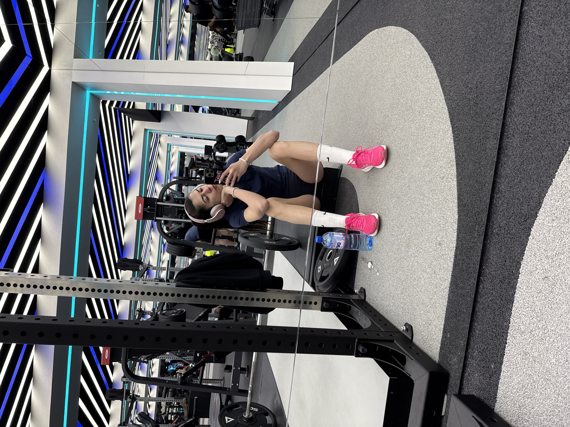A woman is sitting on the floor of a gym in Sofia, Bulgaria, leaning against a weight rack with a water bottle and her phone. She is wearing pink headphones, a dark shirt, dark shorts, white socks, and pink sneakers. A weight plate is next to her and another weight plate sits on the rack. A mirror reflects the gym's interior and an adjacent hallway with alternating black and white stripes and blue lighting. The floor has an abstract grey and black speckled pattern.