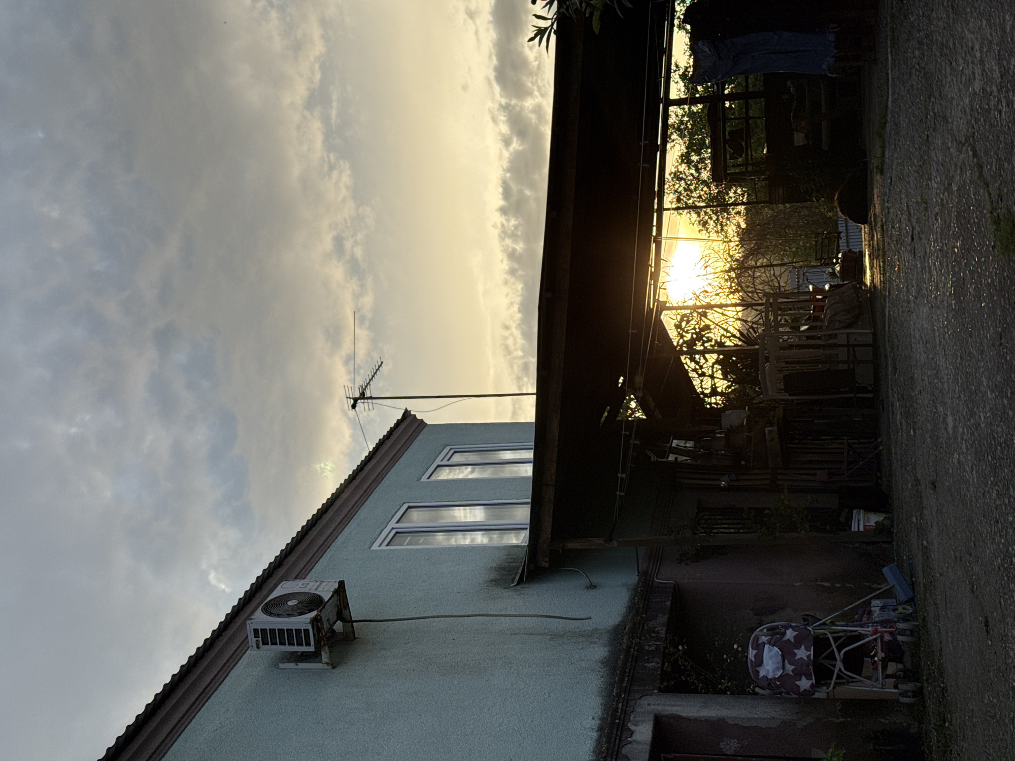 The image depicts a residential building in what may be Sochi, Russia, though this cannot be definitively confirmed from the visual data alone. The building is a two-story structure with a pitched roof and appears to be of older construction. An air conditioning unit is affixed to the exterior wall. An antenna is visible on the roof. The sky is overcast with the sun partially obscured by clouds, casting a muted light on the scene. Adjacent to the building is a covered area filled with various objects and vegetation, creating a cluttered appearance. A stroller is visible near the base of the building. The presence of a concrete pathway suggests a yard or outdoor space. The image is rotated counterclockwise from its presumed original orientation. No people are directly visible.