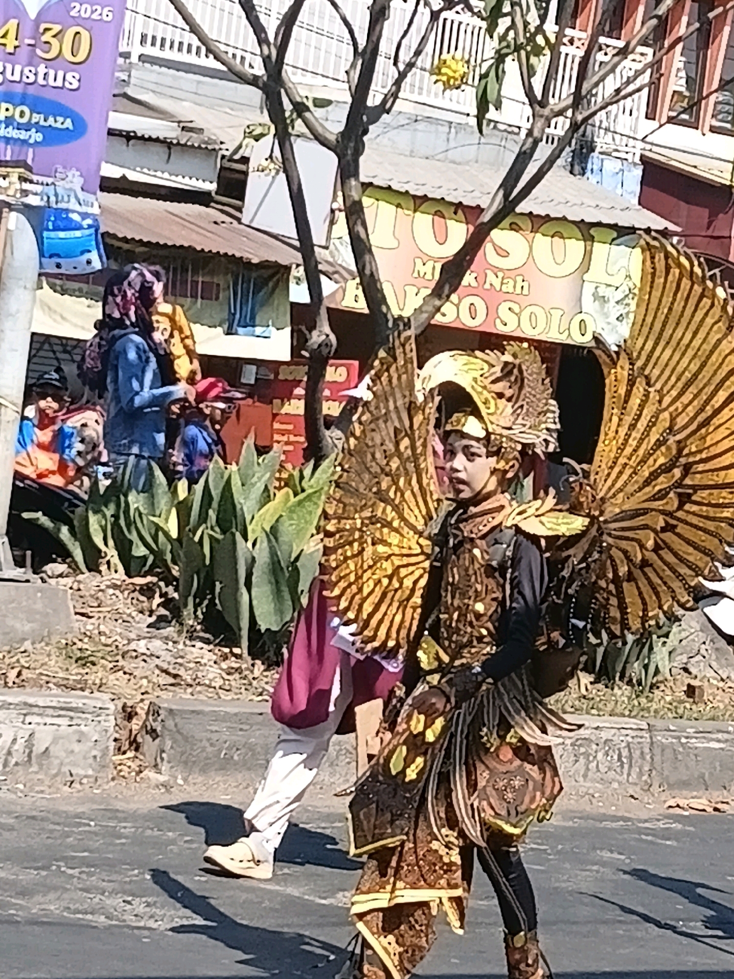 The image depicts a street scene in Wadungasri, Indonesia, during what appears to be a parade or festival. A child dressed in an elaborate, golden bird costume with large wings is walking on a paved street. The costume features intricate patterns and feather-like details. Behind the child, other individuals are visible, some in casual clothing and others in traditional attire, suggesting a cultural event. In the background, there are buildings with corrugated metal roofs, stalls or shops with banners, and lush green foliage, indicating an urban or semi-urban environment. The lighting suggests it is daytime, with the sun casting shadows on the street. One banner in the background advertises "GOPLAZA" and displays dates "2026" and "4-30 Agustus", suggesting a specific event or promotion related to this location and time. Another banner displays "TO SOLO" and "MAS NAH GESO SOLO". The overall atmosphere is lively and celebratory.