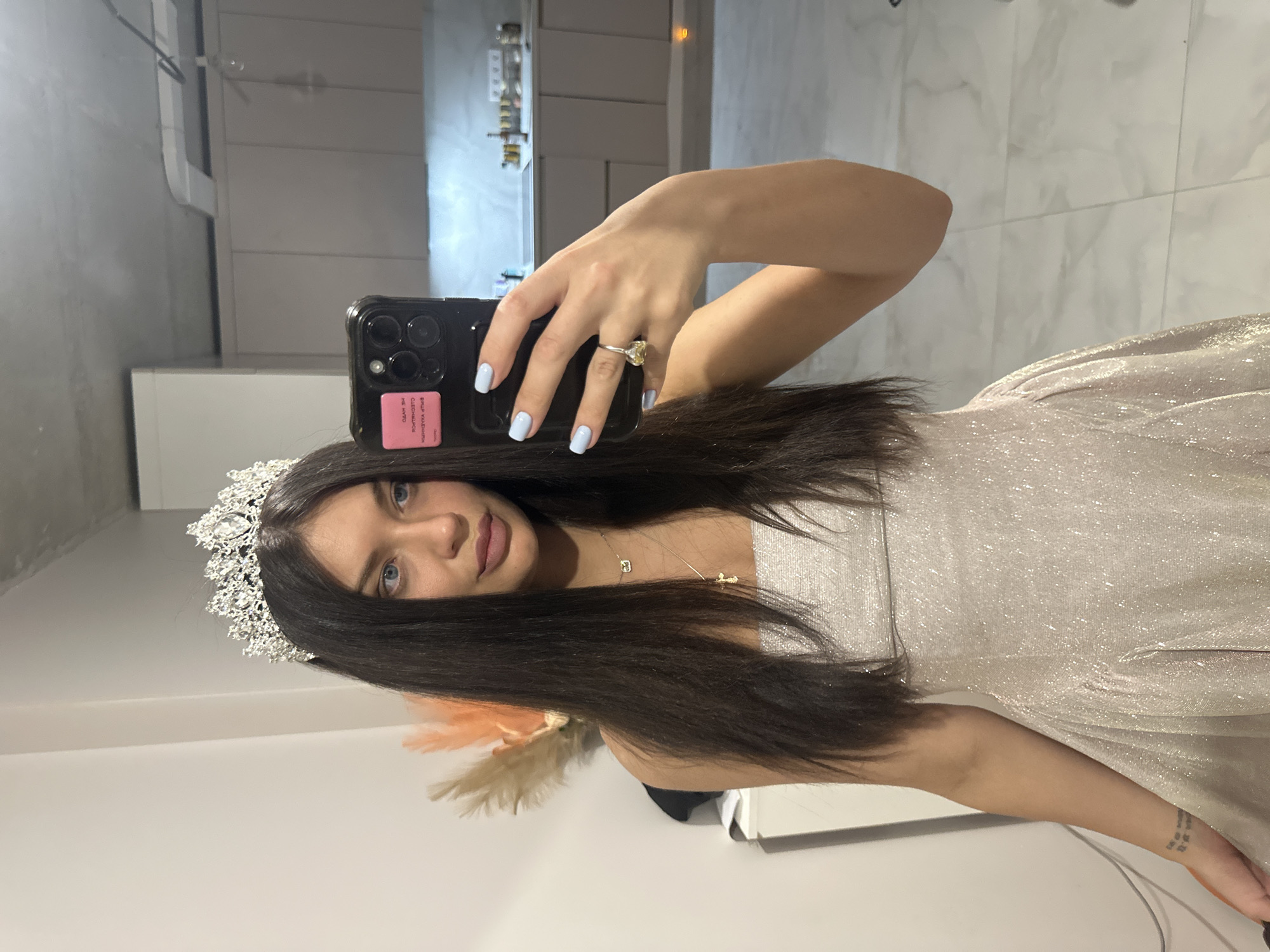 A young woman is depicted taking a mirror selfie, captured indoors in a sleek, modern setting. She has long, dark hair, blue eyes, and a neutral expression as she looks directly into the phone/mirror. She is wearing a shimmering, light-colored (possibly silver or champagne) dress and a prominent, ornate silver tiara on her head.
Her right hand, adorned with light blue nail polish and a ring featuring a large, yellowish gemstone, holds a black smartphone. The phone case has multiple camera lenses and a pink sticker on the back. The text on this sticker is reversed due to the mirror reflection and appears to be in Cyrillic. Around her neck, she wears a delicate necklace with two small charms, one possibly a letter or initial, and the other resembling a cross or a similar symbol. A small, multi-lined tattoo is visible on her left wrist, though the text is too small to decipher.
The background, visible as a reflection in the mirror, suggests a contemporary home interior with light-colored walls, cabinets, and possibly a countertop, indicative of a kitchen or living area. Hints of decorative pampas grass or similar feathery decor are also visible. The event is a personal photo-taking session, likely in the evening or during the day with artificial lighting. The location is Khimki, Russia.