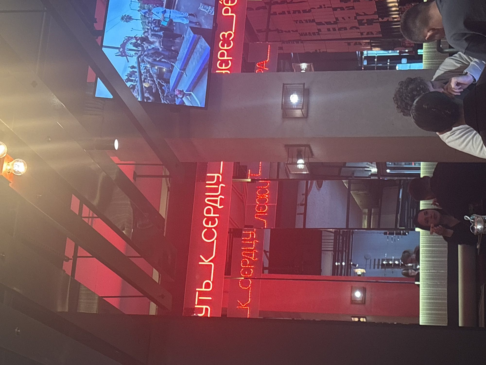 Here is a description of the image:
The image shows an interior view of a restaurant located in Saint Petersburg, Russia. Red neon signs featuring Cyrillic text are a prominent feature of the decor. Patrons are seated at tables in the restaurant, some of whom appear to be engaged in conversation or enjoying a meal. A television screen displays images in the upper part of the frame. The lighting inside the restaurant appears dim, with some light fixtures providing localized illumination. The architecture features metallic support structures and decorative lighting.