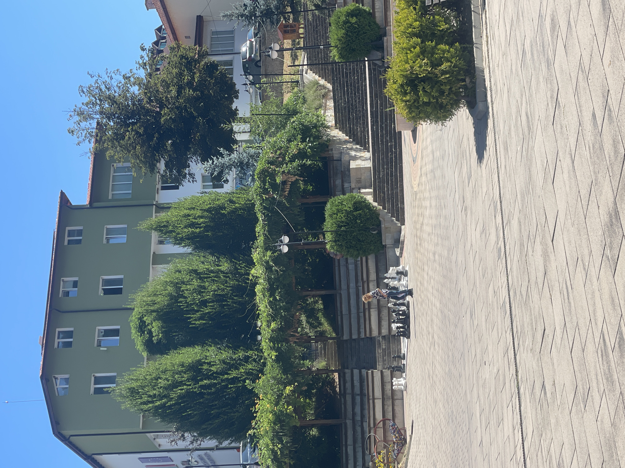 This image depicts a public square in Banite, Bulgaria, on a sunny day. In the foreground, a large outdoor chess set is laid out on the paved ground, with both black and white pieces arranged on the board. A lone individual, wearing a patterned shirt and pants, is walking near the chess set.
Behind the chess set, there are tiered stone seating areas and staircases leading upwards. Lush green trees and shrubbery are strategically placed throughout the scene, providing shade and visual appeal. In the background, a multi-story building painted in green and white stands against a clear blue sky. Other buildings and trees are visible further in the distance, suggesting a typical urban or town environment. The overall scene conveys a sense of leisure and public recreation.