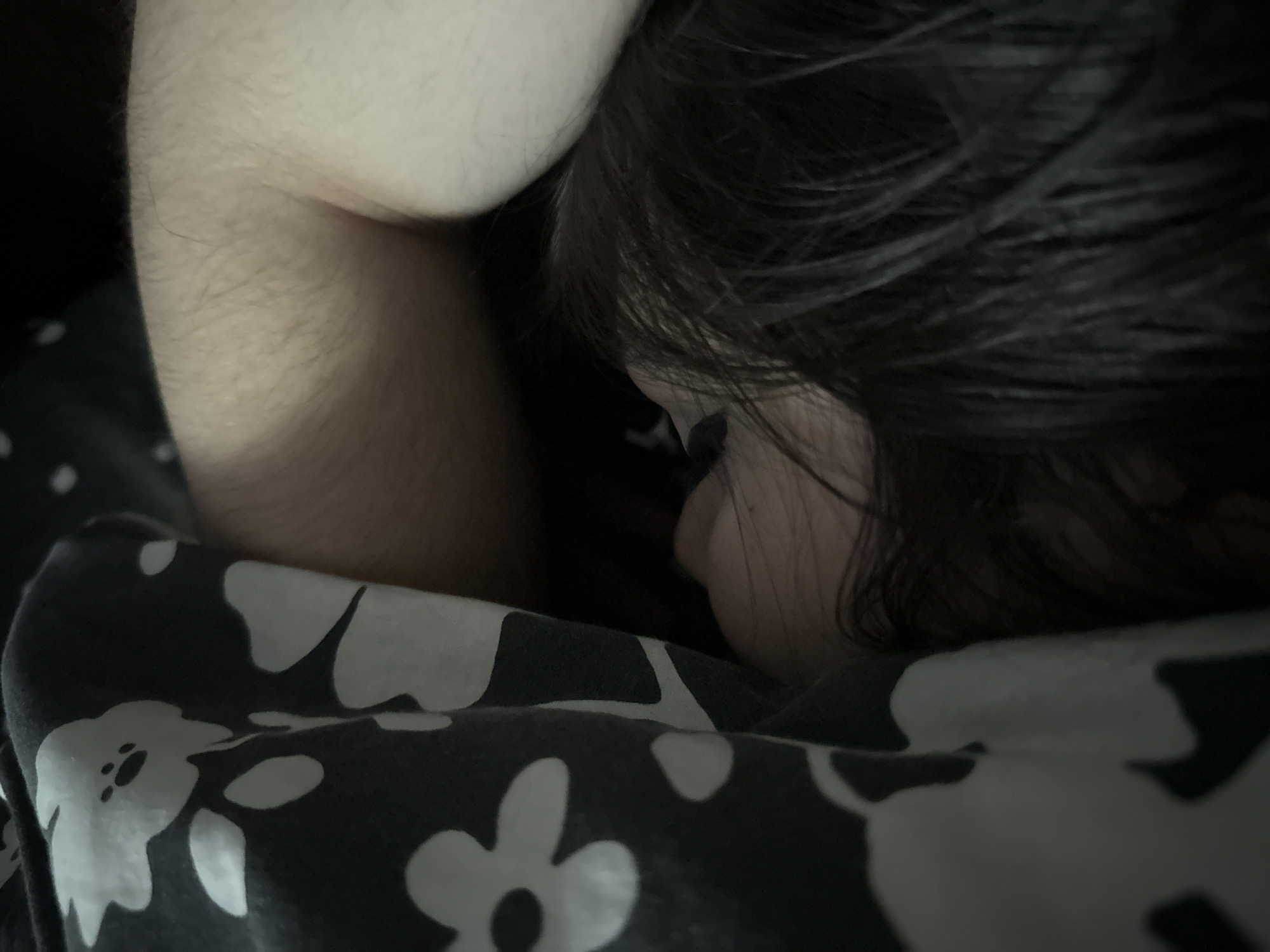 The image presents a close-up, dimly lit scene of a person, likely a child or young adult, sleeping or resting peacefully. The individual is partially visible, nestled under a dark-colored blanket or sheet with a prominent white floral pattern. Their dark hair is visible on the right side of the frame, partially obscuring the face, with only the forehead, eye area, and the side of the nose discernible. An arm or shoulder, covered in fine hair, is positioned near the top left of the frame.
The setting appears to be indoors, possibly a bed or a cozy corner in a room. The low-light conditions suggest it could be nighttime, early morning, or a room with subdued lighting, contributing to a tranquil and intimate atmosphere. No specific location cues are visible within the image, but the provided context places the scene in Blagoevgrad, Bulgaria. The person's posture conveys a sense of comfort and deep relaxation. There is no visible text in the image.