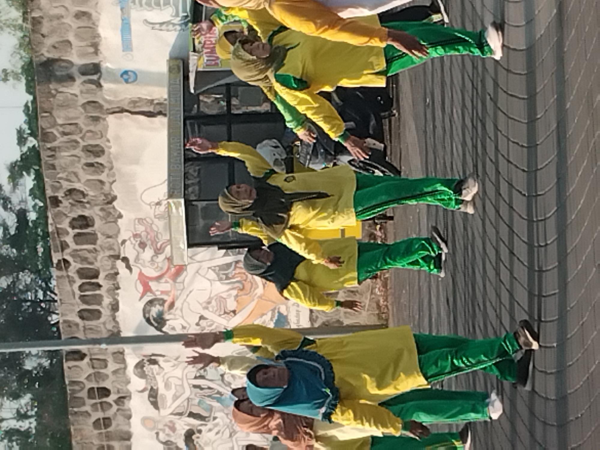 The image depicts a group of women engaged in an outdoor exercise activity, likely a form of aerobic dance or stretching. They are dressed in matching bright yellow long-sleeved shirts with green trim and green athletic pants. Most of the women are wearing hijabs in various colors, including brown, teal, and light green. Their arms are raised in unison as they perform synchronized movements.
The background features a textured stone wall and a mural painted on a lighter surface. The mural appears to depict stylized figures and perhaps mythological or traditional imagery, with elements of red, black, and white. Below the mural, there is a bus or vehicle with signage. One sign reads "ROTI BAKAR TUAN MUDA" and another appears to be for "LUMPI...". There is also a streetlight pole on the left side of the image. The ground is paved with interlocking brick pavers that create a patterned surface.
The scene is set in an urban environment, possibly a public square or promenade. The lighting suggests it is daytime, with sunlight casting shadows on the ground. The presence of a motorcycle on the right side of the image further indicates an urban setting. This activity appears to be taking place in the city of Karanganyar, Indonesia, given the cultural attire and the style of signage visible.