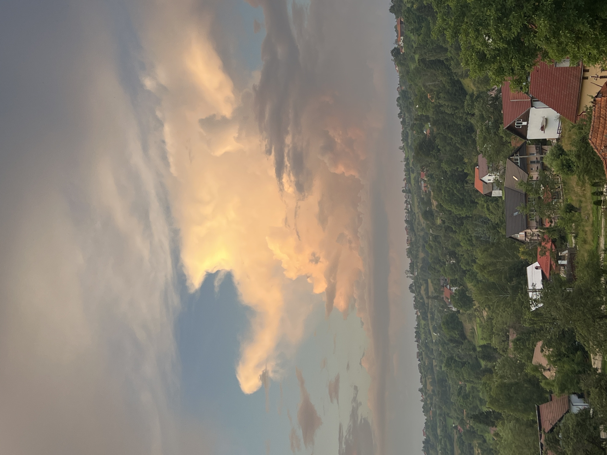 The image depicts a rural landscape under a dramatic sky. Dominating the upper portion of the frame is a large, illuminated cloud formation, tinged with warm, golden hues from what appears to be the setting sun. This prominent cloud is set against a backdrop of softer, more diffused grey and pale blue sky. Below the sky, a rolling green hillside is covered in a dense canopy of trees. Scattered amongst the trees are numerous houses, varying in size and architectural style, with red tile roofs being a common feature. The houses are nestled into the contours of the hill, suggesting a small village or scattered rural dwellings. The foreground features more closely situated houses and vegetation, hinting at the edge of a settlement. The overall impression is one of a tranquil, scenic countryside at dusk, with the striking cloud formation adding a dynamic element to the scene.