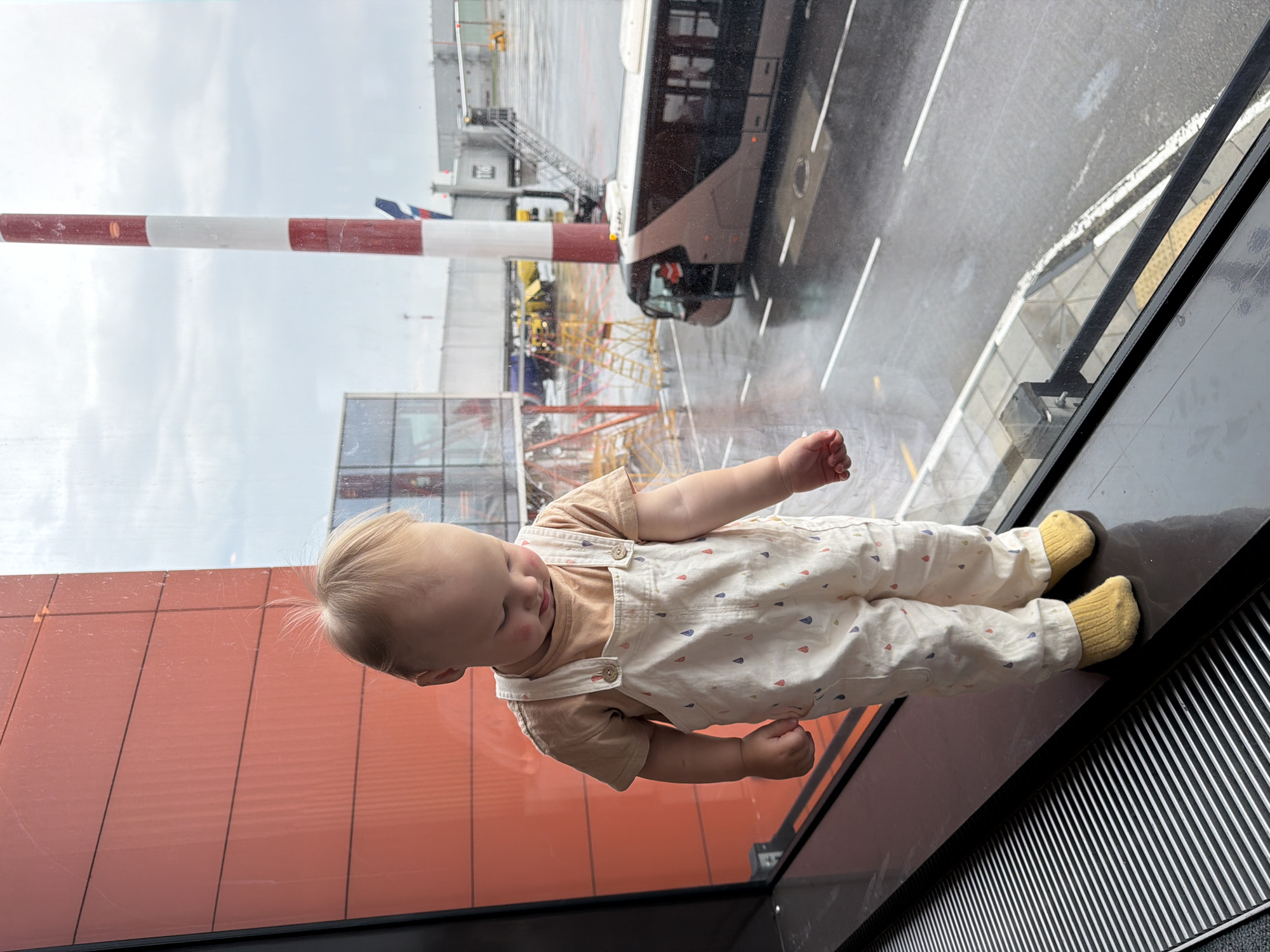 A young child stands on a dark, reflective surface next to a large window. The child is wearing a light-colored outfit with a pattern of colorful drops, and yellow socks. Their head is bowed, and their gaze is directed downward. Outside the window, an airport tarmac is visible under a cloudy sky. A bus with its door open is parked on the wet asphalt, and a red and white striped pole stands nearby. In the background, there are airport jet bridges and other airport structures. The overall setting appears to be an airport in Khimki, Russia, possibly Terminal B of Sheremetyevo International Airport, given the architecture and typical airport elements.
