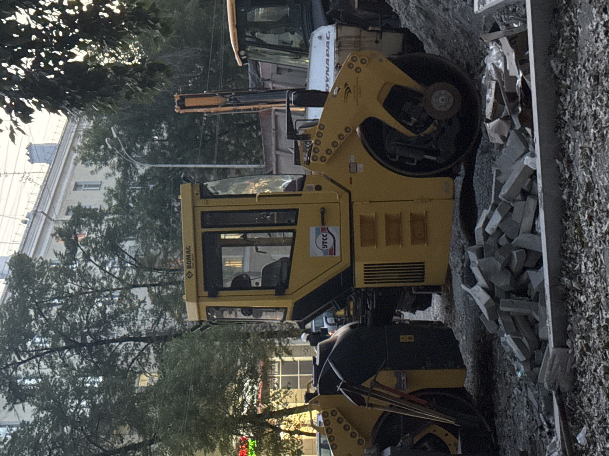 The image depicts a street scene in Yekaterinburg, Russia, likely during daytime given the lighting. A large yellow Bomag brand soil compactor is prominently positioned in the foreground, with its drum visible on the left side. The compactor features a glass cab and has the "UTCC" logo on its side. Behind the compactor, parts of other heavy machinery are visible, including what appears to be a Dynapac brand loader or backhoe. The ground is covered with gravel and debris, including a pile of broken bricks, suggesting ongoing construction or roadwork. In the background, trees line the street, and a multi-story building with windows and an unusual roof feature is visible. A streetlamp is also present. A person wearing a white helmet is visible in the middle ground, near the compactor, though their actions are not clear. Another person is visible through the window of the compactor. The overall impression is one of an active construction site within an urban environment.