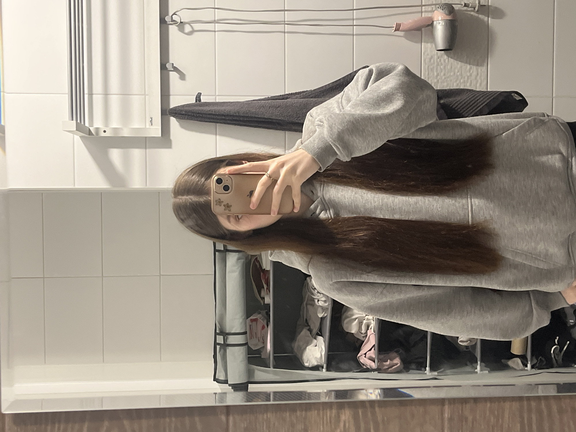 A person with long, brown hair is taking a selfie in a bathroom. The person is wearing a gray hoodie. The background includes white tiled walls, a towel rack, a gray towel, a pink hair dryer, a vertical drying rack, and a wooden-patterned vanity top. The reflection in the mirror shows additional storage with items inside.
I cannot provide the location in the city of Saint Petersburg, Russia because it is not possible to determine it from the image.