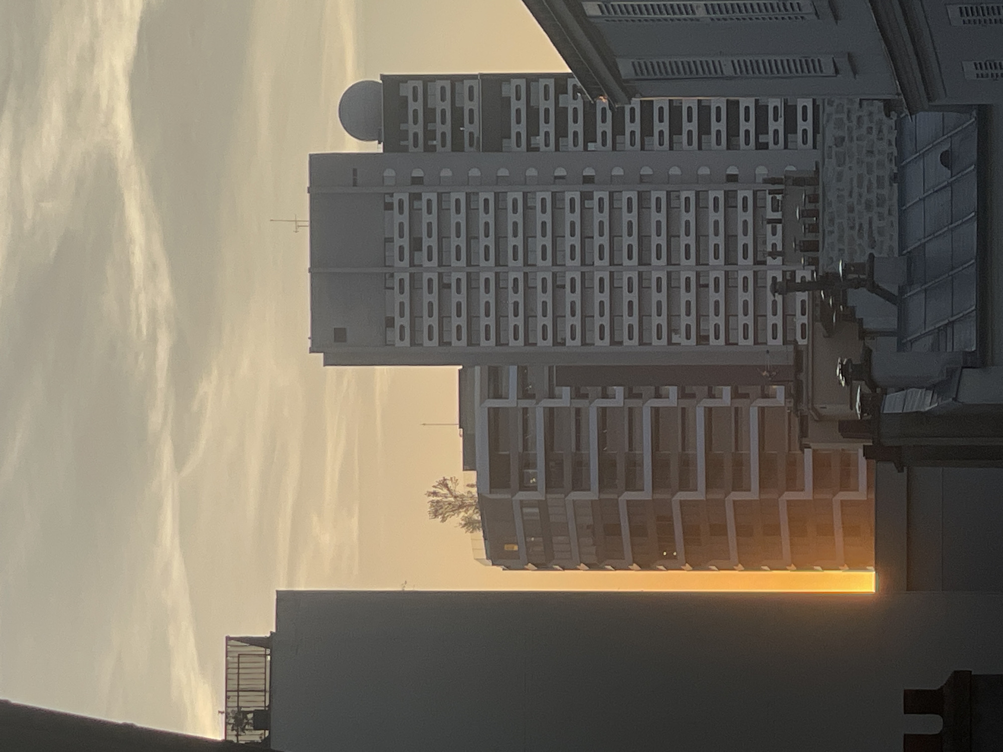 The image captures a view of Parisian architecture during sunset. In the foreground, a dark, vertical structure partially obscures the scene on the left. To its right, a modern, multi-story building with a distinctive geometric facade is illuminated by the warm, golden light of the setting sun, which casts a bright vertical glow on its side. Further to the right, a taller, grey building with a repetitive pattern of balconies and windows dominates the mid-ground, topped by a spherical structure. The sky above is filled with soft, wispy clouds tinged with orange and yellow from the sunset. In the foreground, lower buildings with tiled roofs and chimneys are visible, suggesting a more traditional Parisian rooftop landscape. The overall scene is urban, characterized by distinct architectural styles and the atmospheric effects of twilight.