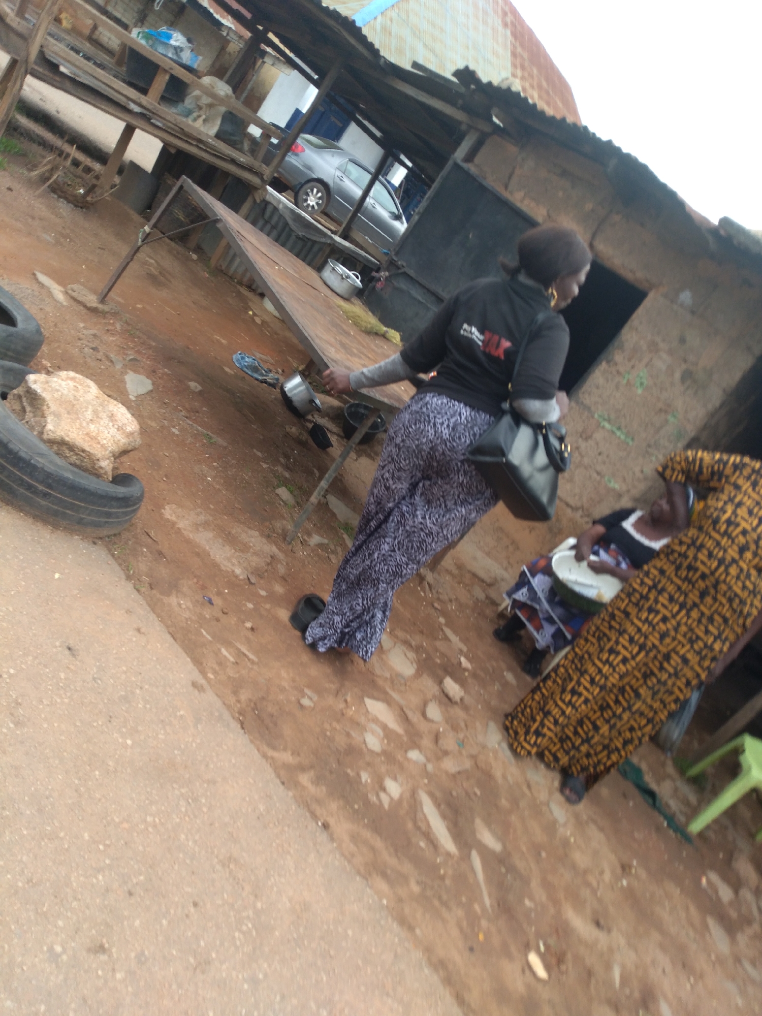 The image depicts an outdoor scene, likely in Jos, Nigeria, characterized by a rustic market or roadside stall environment. In the foreground, a person in a black t-shirt with a red logo and patterned trousers walks away from the camera, carrying a black handbag. Behind them, another person sits, wearing a black and white top and a blue and red patterned wrap skirt, holding a white bowl. To the right, a person in a yellow and black patterned garment is partially visible. The ground is a mix of dirt and rough pavement. Several objects are scattered around, including tires, a large rock, metal containers, and what appears to be a portable table or stall. In the background, a silver car is parked near a structure with a corrugated iron roof and wooden supports, suggesting a simple, utilitarian setting. The overall impression is one of a busy, informal commercial area.