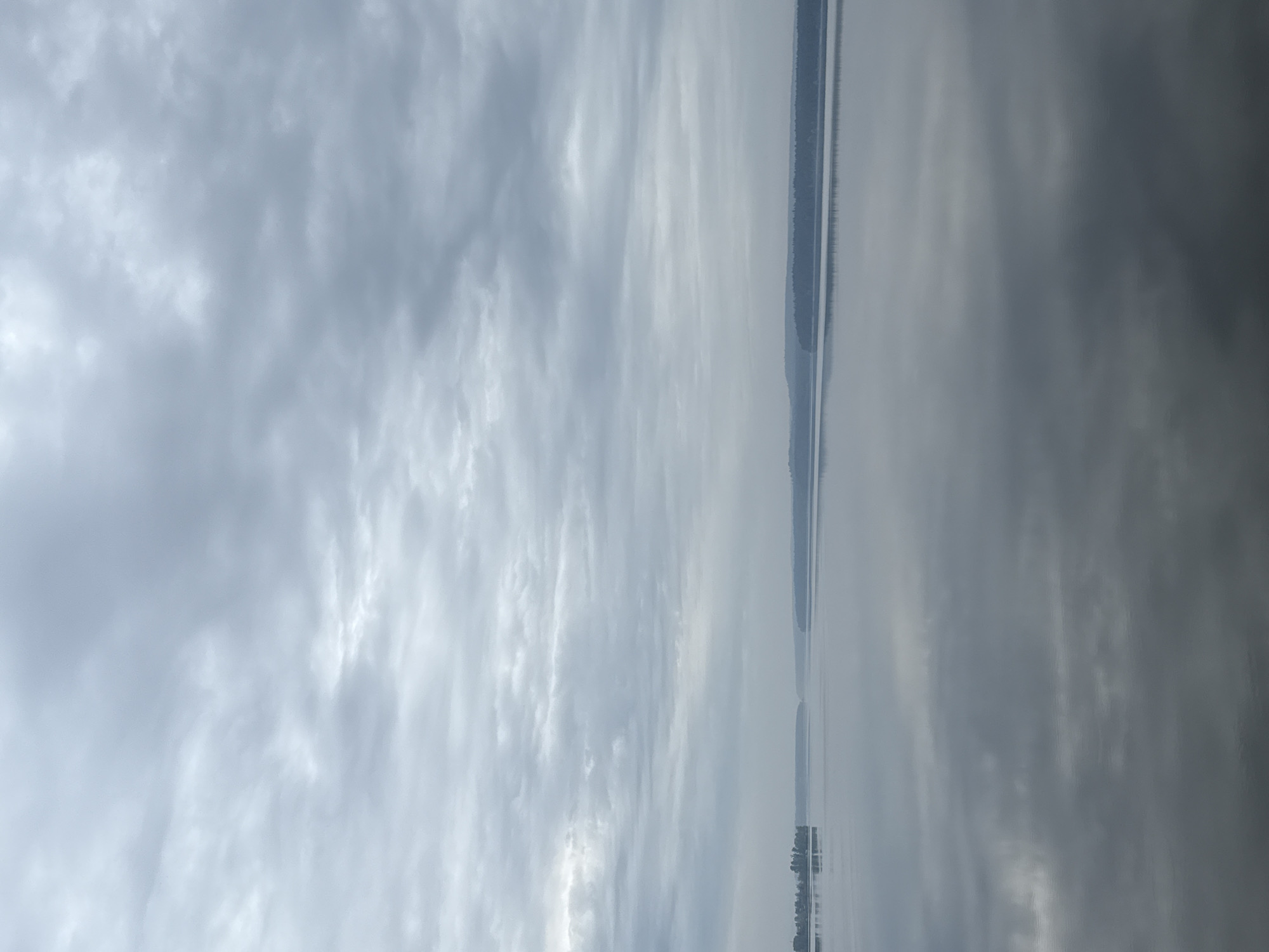 The image presents a view of the Votkinsk Reservoir in Votkinsk, Russia, under an overcast sky. The surface of the water reflects the cloudy sky above, blurring the distinction between the sky and the water. A thin strip of land is visible on the opposite shore, appearing dark against the pale sky and water. The composition of the image is dominated by the sky and water, with the land acting as a subtle horizon line.