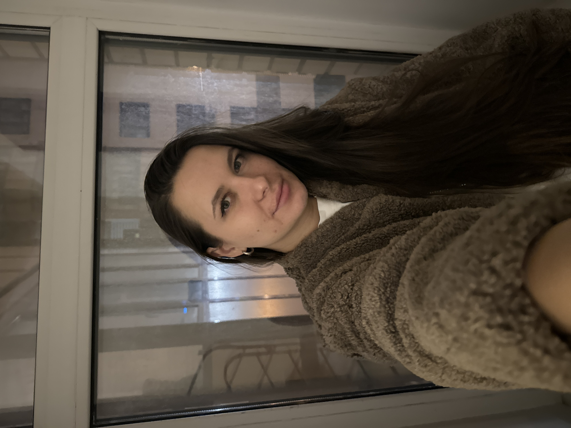 A woman with long brown hair is indoors, wearing a brown, fluffy bathrobe. The background is a window showing an indistinct, blurred cityscape, possibly containing buildings and street lighting. The location is likely Akulovo, Kubinka, Russia, based on the prompt.