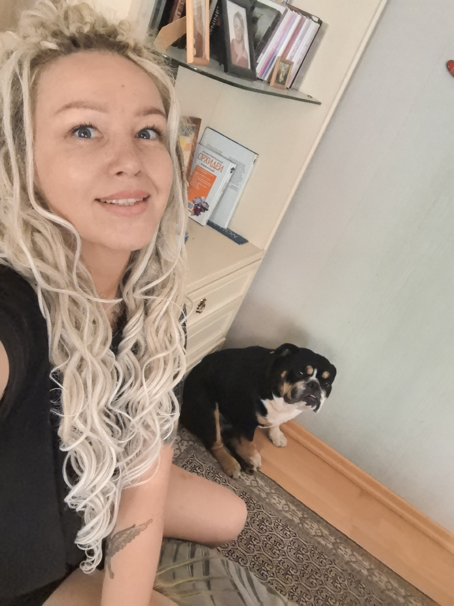 A woman with blonde dreadlocks and a black shirt takes a selfie with an English bulldog. The woman is smiling, and the dog is looking at the camera. They are in a room with a dresser, a shelf with books and picture frames, and a wall. The floor is carpeted. The setting appears to be domestic. There is no information in the image that indicates the location is Nizhny Novgorod, Russia.