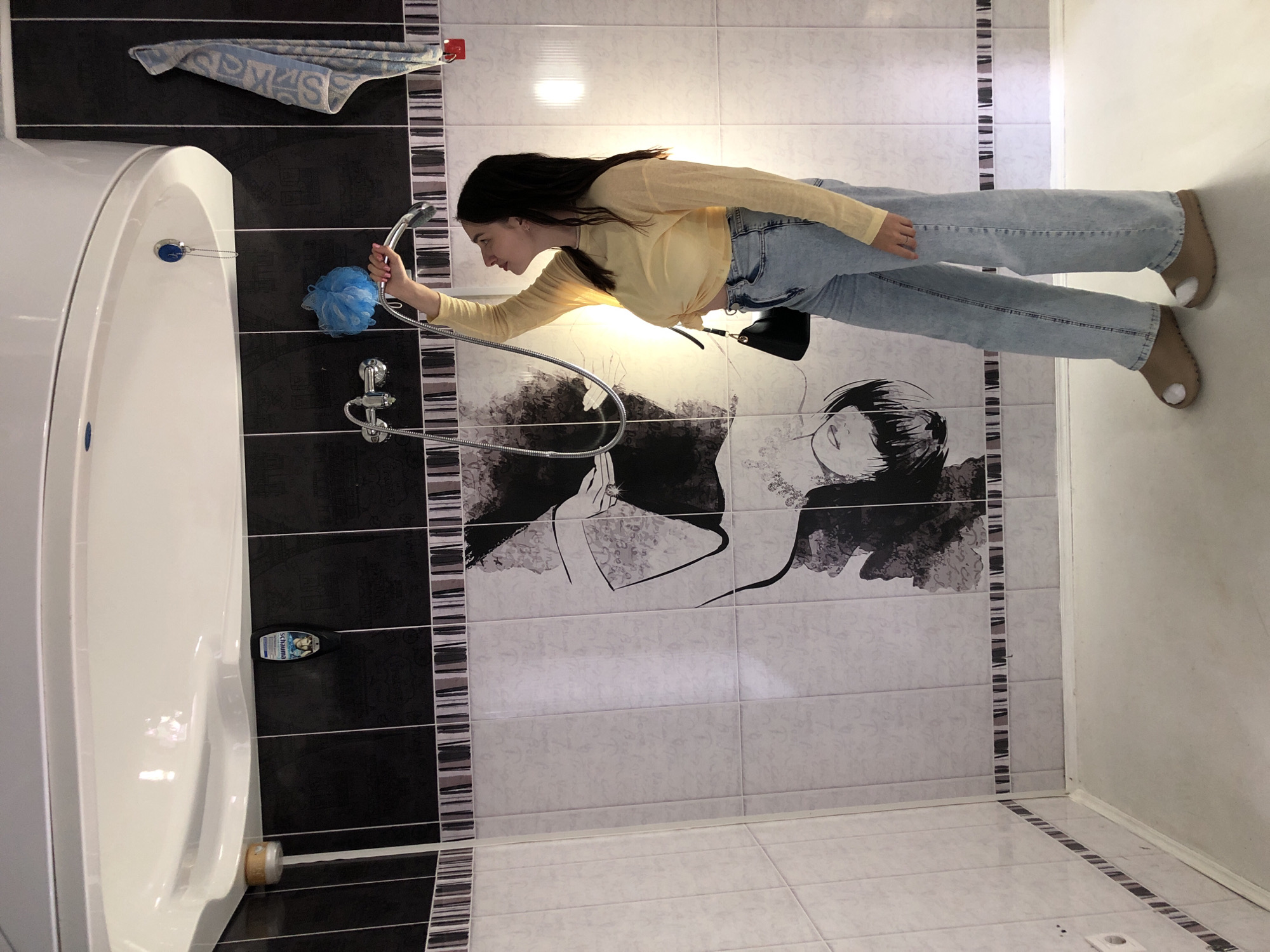 A young woman with dark hair is standing in a bathroom, holding a shower head. She is wearing a yellow long-sleeved shirt and light blue jeans. The bathroom has black and white tiled walls with a decorative pattern. A white bathtub is visible on the left side of the image. A blue bath sponge hangs on the wall near the bathtub. A towel is draped over a bar above the bathtub. The floor is tiled with white and black patterned tiles. The scene appears to be indoors, likely in a residential bathroom. The lighting suggests it is daytime. There is no visible text in the image.