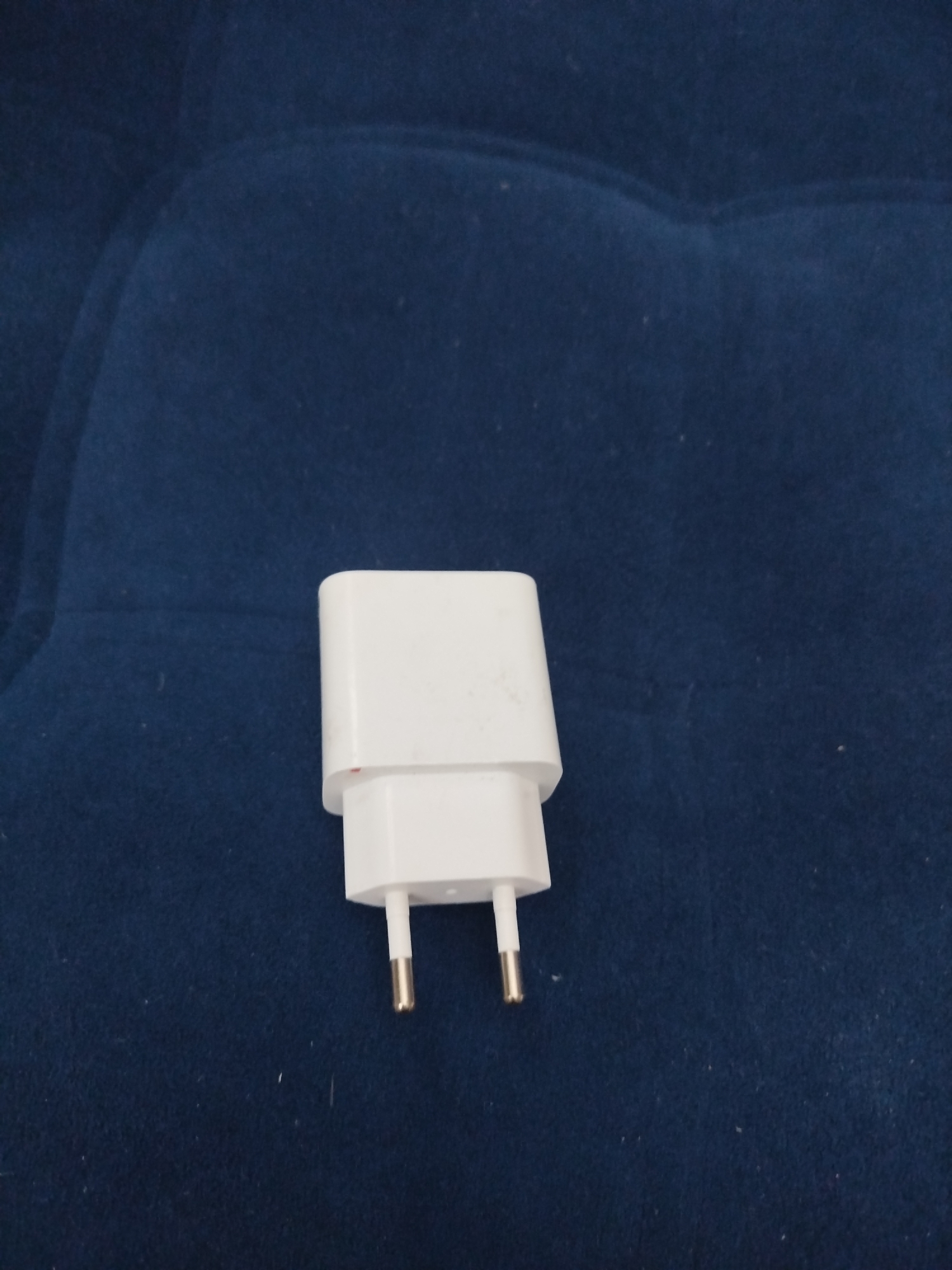 A white power adapter is positioned on a dark blue, textured surface. The adapter has two metal prongs extending downwards. The surface appears to be fabric, possibly upholstery or a blanket, with a slightly rippled texture. The image is a close-up shot, focusing primarily on the adapter. There is no indication of the location being Zhigulyovsk, Russia.