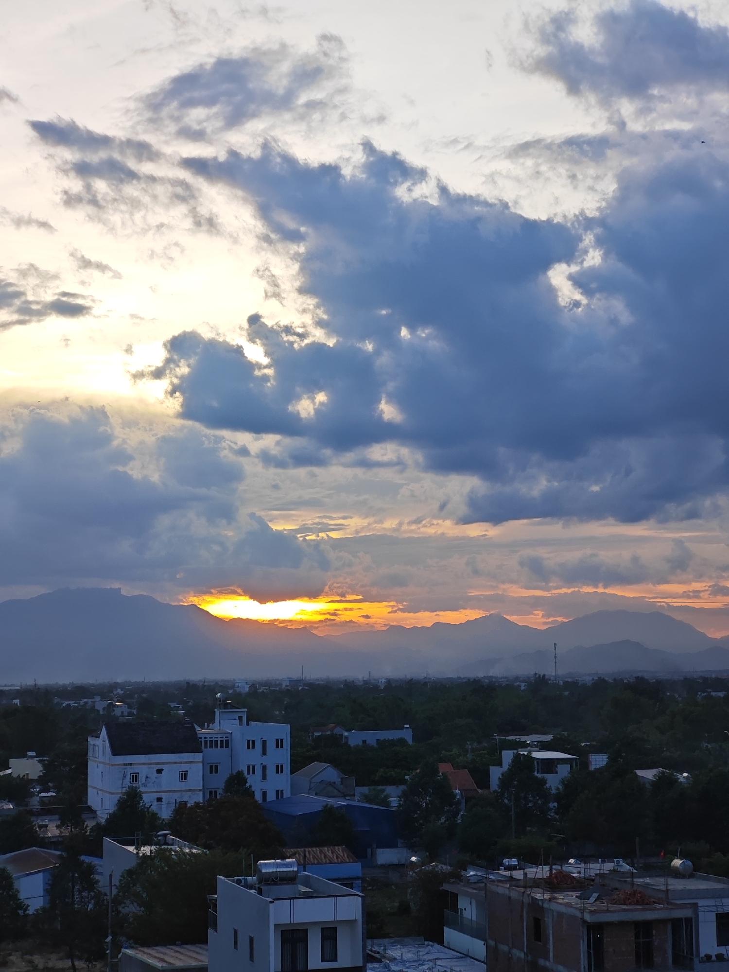 The image captures a sunset over a cityscape, likely in the Cam Le district of Da Nang, Vietnam, given the mountainous backdrop and urban development. A cluster of buildings, including a prominent white structure with multiple windows and a flat roof, dominate the foreground. Other structures, some under construction, are also visible. Dense foliage, including trees, surrounds the buildings, suggesting a semi-urban or suburban environment. In the distance, a mountain range is silhouetted against the vibrant sky, with the sun peeking through dramatic clouds. The sunset casts a warm, orange glow across the scene, illuminating parts of the sky and the tops of the mountains. The overall atmosphere is one of a serene evening transition in a Vietnamese urban landscape.
