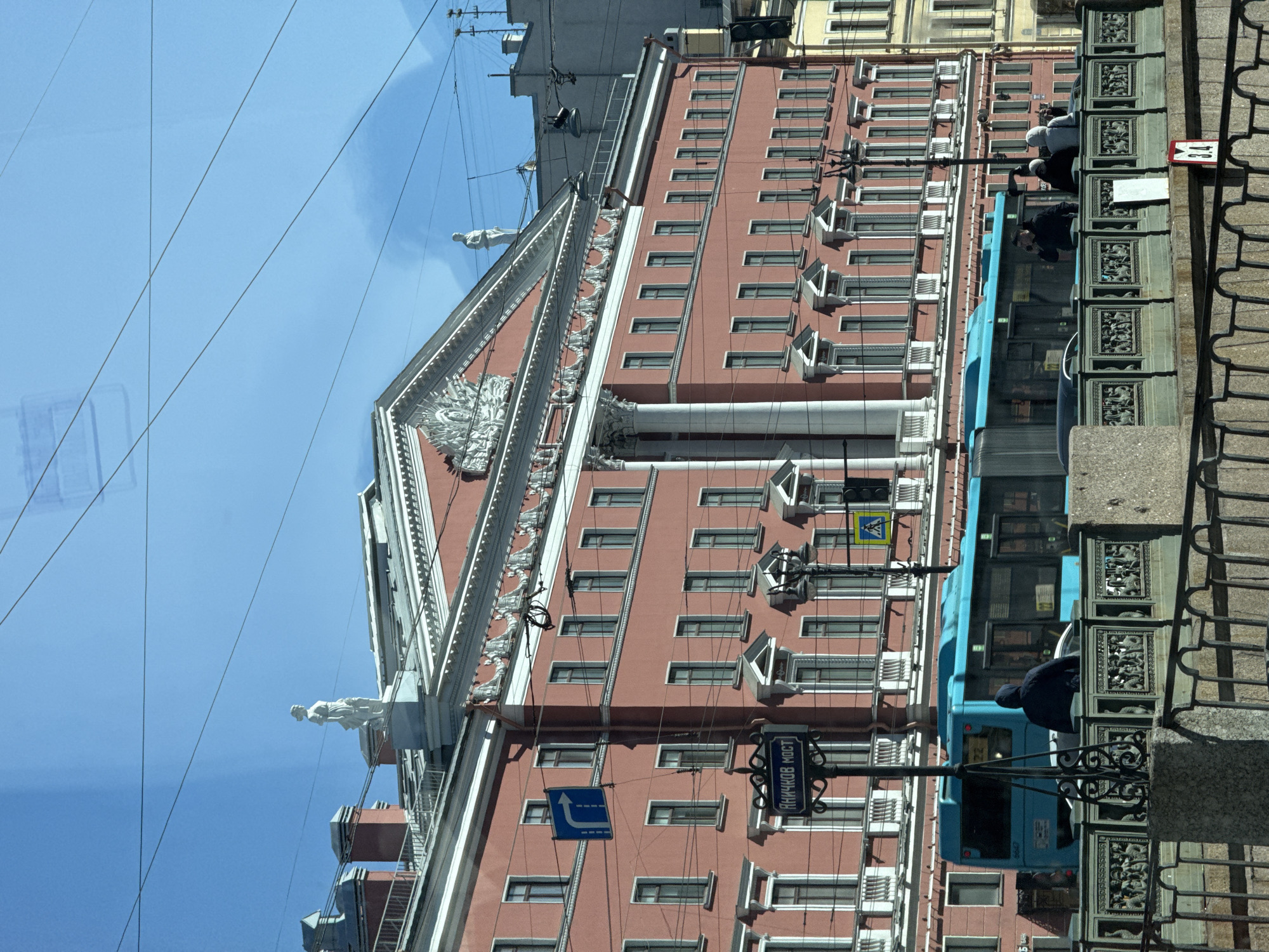 The image shows a building in Saint Petersburg, Russia, viewed from a moving vehicle. The building is tall and pink, with multiple windows and decorative architectural elements, including a statue on the roof and ornate trim. A street sign reading "ЯНКУКОВ МОСТ" (Anichkov Bridge) is visible on the building's facade. A blue trolley bus is passing by, and a wrought-iron fence lines the street. The sky is blue and clear.