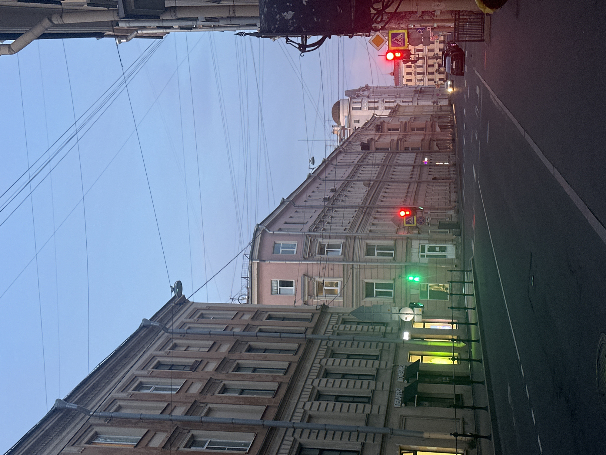 The image depicts an urban street scene at dusk in a city, likely European, given the architecture. On the left, a multi-story building with a beige facade features numerous windows and decorative architectural elements. Below, a storefront with illuminated windows displays signage that appears to be in Cyrillic script, possibly indicating a bakery and coffee shop. A black metal fence runs along the sidewalk in front of the building. Across the street, a row of similar multi-story buildings with varied facades and window arrangements lines the right side. A traffic light is visible, currently showing green, and further down the street, another traffic light is red, impacting traffic flow. Several cars are present on the road, including a black SUV and a white sedan. Overhead, a network of power lines crisscrosses the sky, and streetlights are mounted on poles. The sky is a pale blue transitioning to a faint pinkish hue near the horizon, suggesting the twilight hour. The road surface is asphalt with white lane markings.
