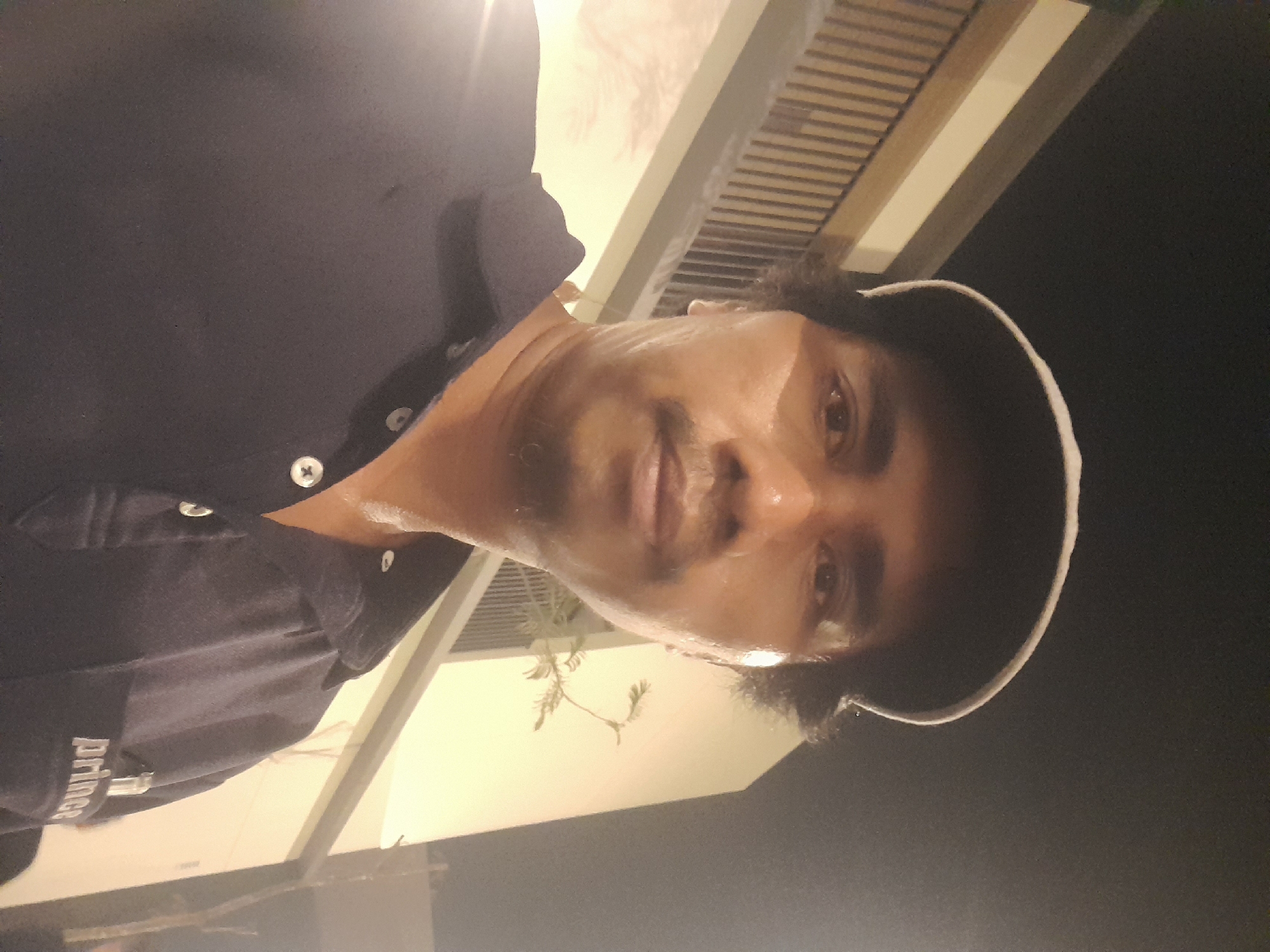 A man wearing a dark blue polo shirt and a dark baseball cap is pictured in a selfie. He has dark, curly hair and a mustache. The background suggests an outdoor setting at night, with a building that has a balcony and railing visible. The man appears to be sweating. The image is a close-up, focusing on the man's face and upper body. The location in Kragilan, Indonesia is noted.