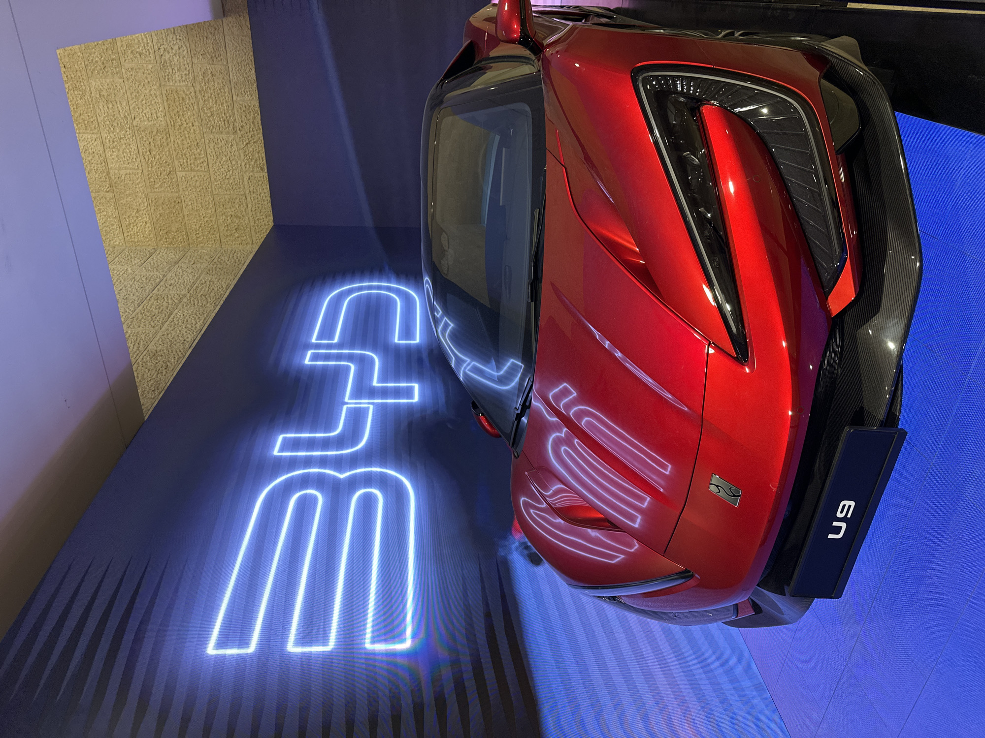 The image depicts a red car, possibly a sports car, positioned on a blue surface with illuminated lettering, which appears to be part of an exhibit or display. The car's rear is in focus, showing details like its taillights and a black panel with the inscription "U9". The setting includes a textured beige wall on the left side and a blue backdrop that suggests an indoor exhibition space. While the image provides limited contextual information, it does not offer evidence to verify that this is in the city of Tel Aviv-Yafo, Israel, or any information about people, actions, or interactions occurring within the scene.