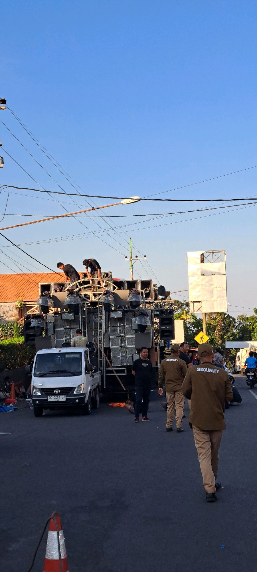 The image depicts an outdoor scene with a large sound system setup on a truck in what appears to be a street in Prigen, Indonesia. The sound system is elaborate, with numerous speakers stacked high on a metal frame. Several individuals are working on the sound system, with two men in black t-shirts seen on top of the structure, appearing to be adjusting or setting it up.
In the foreground, a white pickup truck with the license plate "AG 9456 VG" is parked. Several individuals dressed in khaki uniforms with the word "SECURITY" printed on the back are visible, some standing and observing, while others are walking. One security guard is in the immediate foreground, with his back to the camera.
The environment suggests a public street with power lines overhead and buildings in the background, including a house with an orange roof. The sky is clear and blue, indicating daytime. There are also some traffic cones and other debris scattered on the side of the road. The overall scene suggests preparation for an event that involves a significant sound setup.