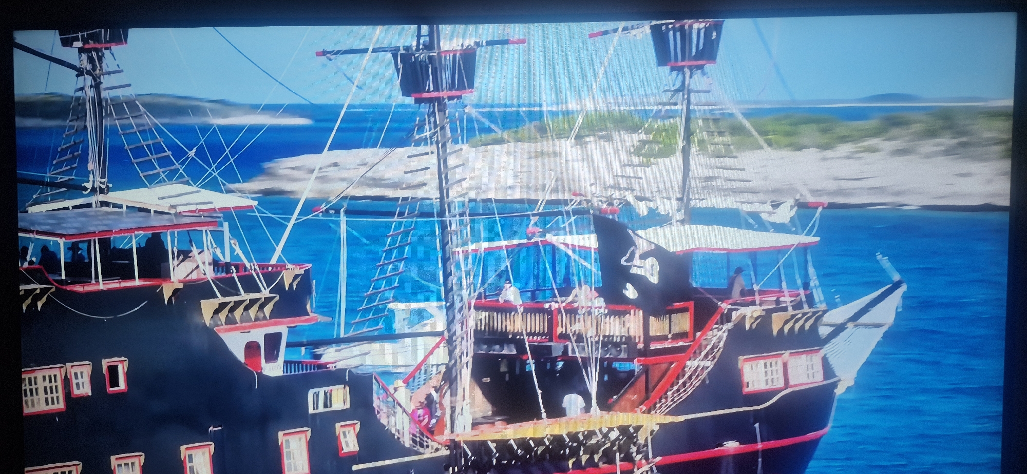 The image shows a large pirate ship sailing on a bright blue sea with islands in the background. The ship has black and red trim, multiple decks, and tall masts with rigging. People are visible on the decks. A pirate flag is flying. The location is unspecified but appears to be a coastal area with clear, blue water.