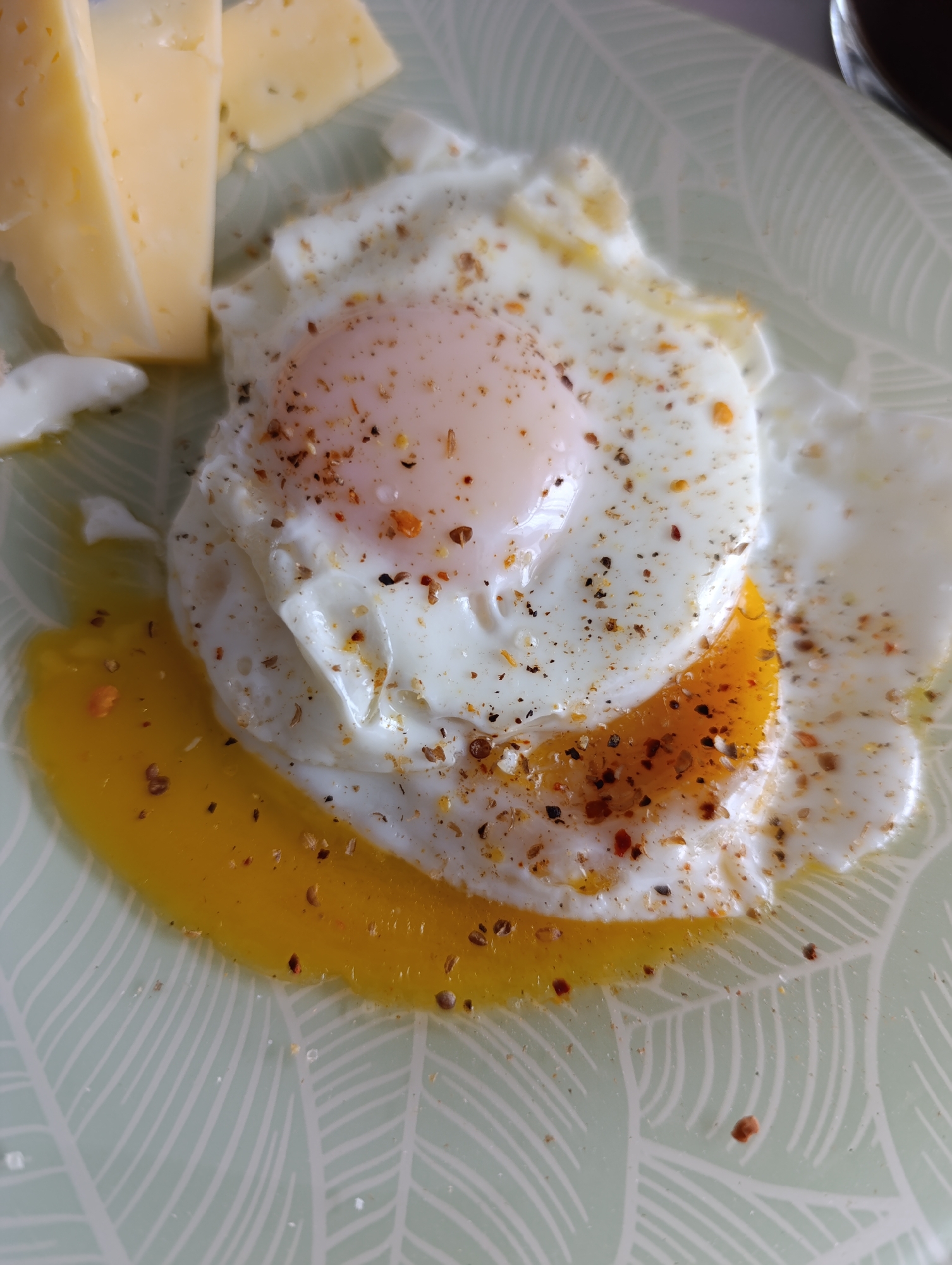 A close-up shot shows a fried egg with a runny yolk, seasoned with pepper and herbs, next to two wedges of cheese on a patterned plate. The plate has a light green background with white leaf-like designs. The egg yolk is dripping onto the plate. The cheese wedges are varying shades of yellow. The environment seems to be a kitchen or dining area. There is no information to suggest a location in the city of Chaikovsky, Russia.