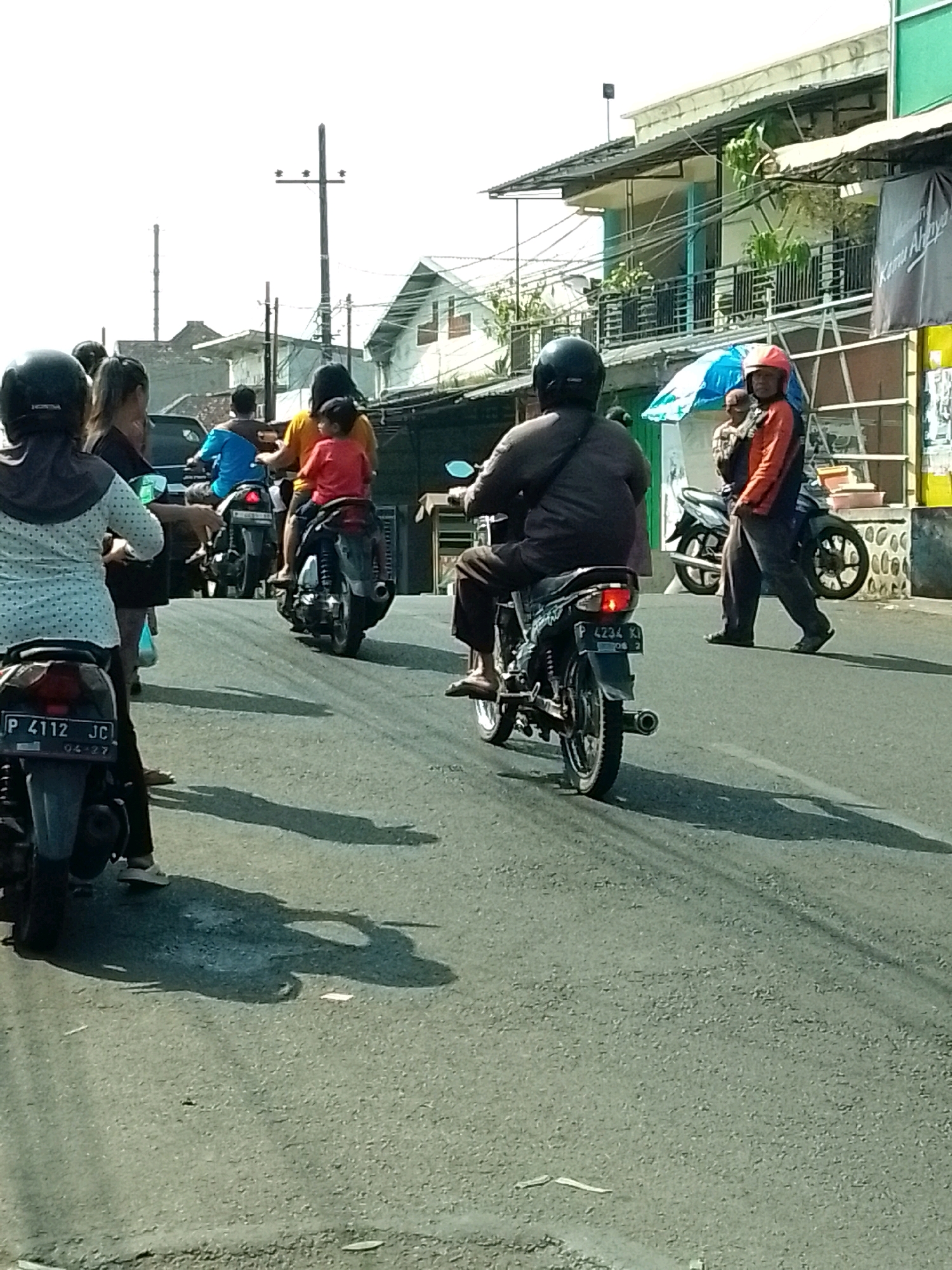 In Jember, Indonesia, a street scene depicts several motorcycles on a paved road under a bright sky. A motorcycle with license plate P 4112 JC is prominently featured in the foreground, facing away from the camera. Behind it, another motorcycle with license plate P 4234 K is also visible. A group of individuals are on or around motorcycles. One person, wearing a dark helmet and jacket, is riding a motorcycle. Another individual, in an orange jacket and dark pants, stands near a motorcycle. Further down the road, a person wearing a blue shirt is on a motorcycle, accompanied by passengers. Buildings with balconies and corrugated roofs line the street. A utility pole with crossbars stands tall. The overall environment suggests a busy street with moderate traffic. Shadows cast by the motorcycles and people indicate it is daytime.