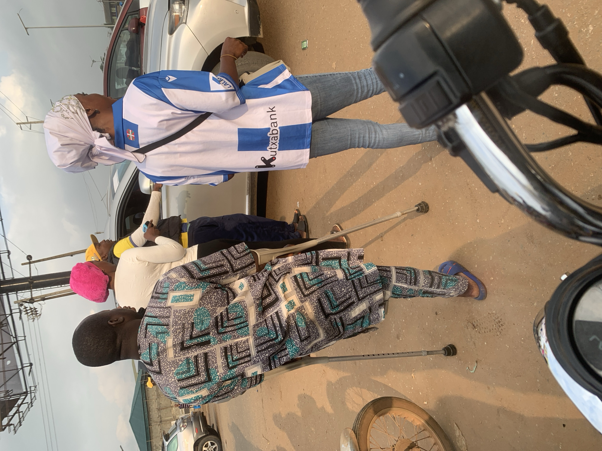 In Shagamu, Nigeria, three people are visible on a dirt road with a light pole and overhead wires in the background. On the left, a man on crutches wears a patterned tunic and blue sandals. In the center, a person in a white shirt and a pink headscarf stands next to another person in a yellow shirt and a yellow cap. To the right, a person wearing a white and blue jersey with "Kutxabank" printed on it and blue jeans is visible. Several cars are parked behind the individuals. The lighting suggests it is either early morning or late afternoon.