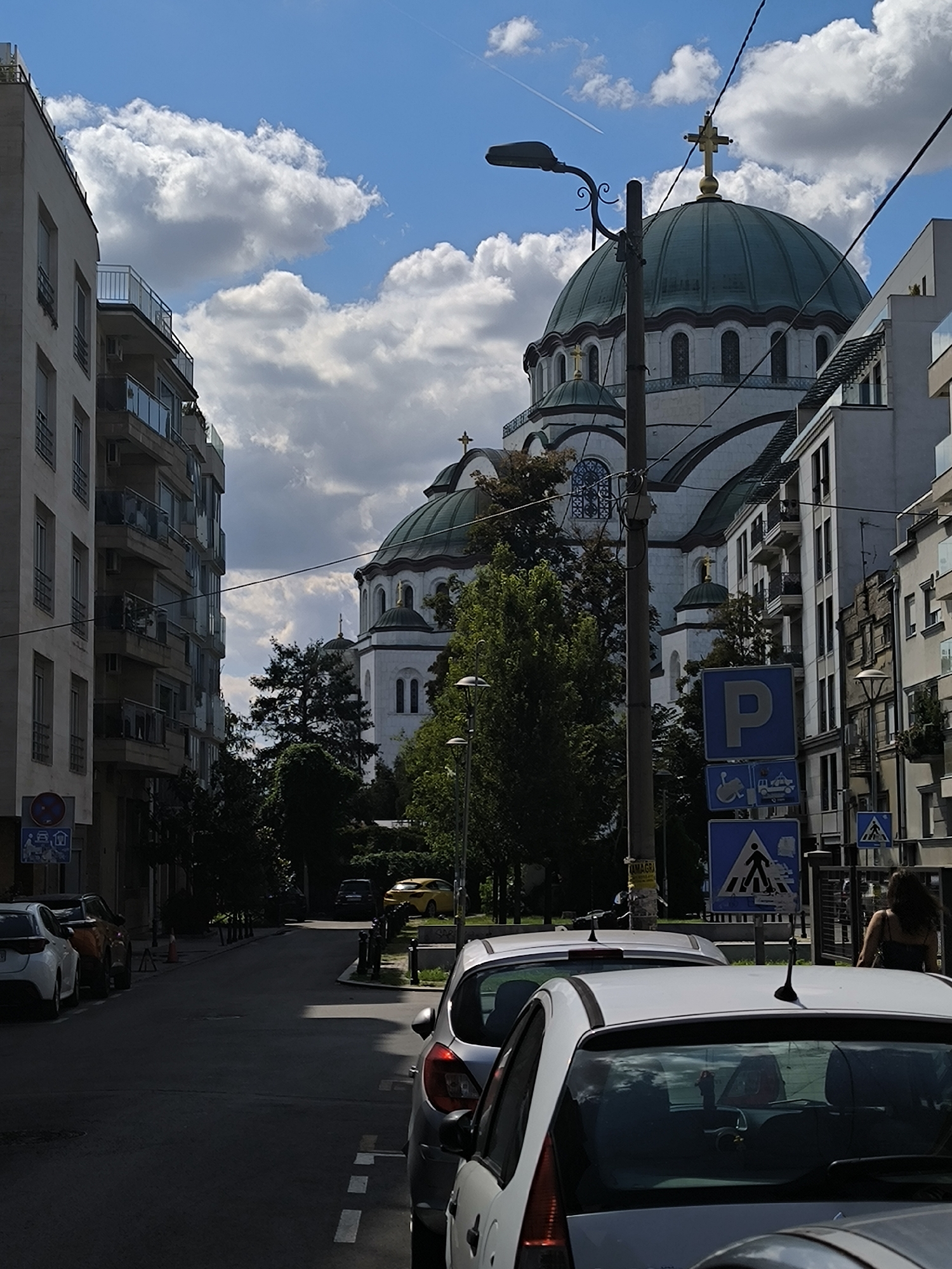 This image depicts a street scene in Belgrade, Serbia, dominated by the large green dome of the Church of Saint Sava. The perspective is from ground level, looking up and down a street lined with modern apartment buildings and trees. The sky is blue with scattered white clouds.
In the foreground, several cars are parked on the side of the street. The closest car is white. Further down the street, a yellow car is visible. There are street signs, including a parking sign and a pedestrian crossing sign.
The Church of Saint Sava is a prominent landmark, with its imposing dome and white facade. It is partially obscured by trees and other buildings. A lamppost is visible in the middle ground, close to the church.
A woman with dark hair is walking away from the camera on the right side of the image, in front of a modern building. The overall impression is of a sunny day in an urban environment, with a mix of residential and religious architecture.