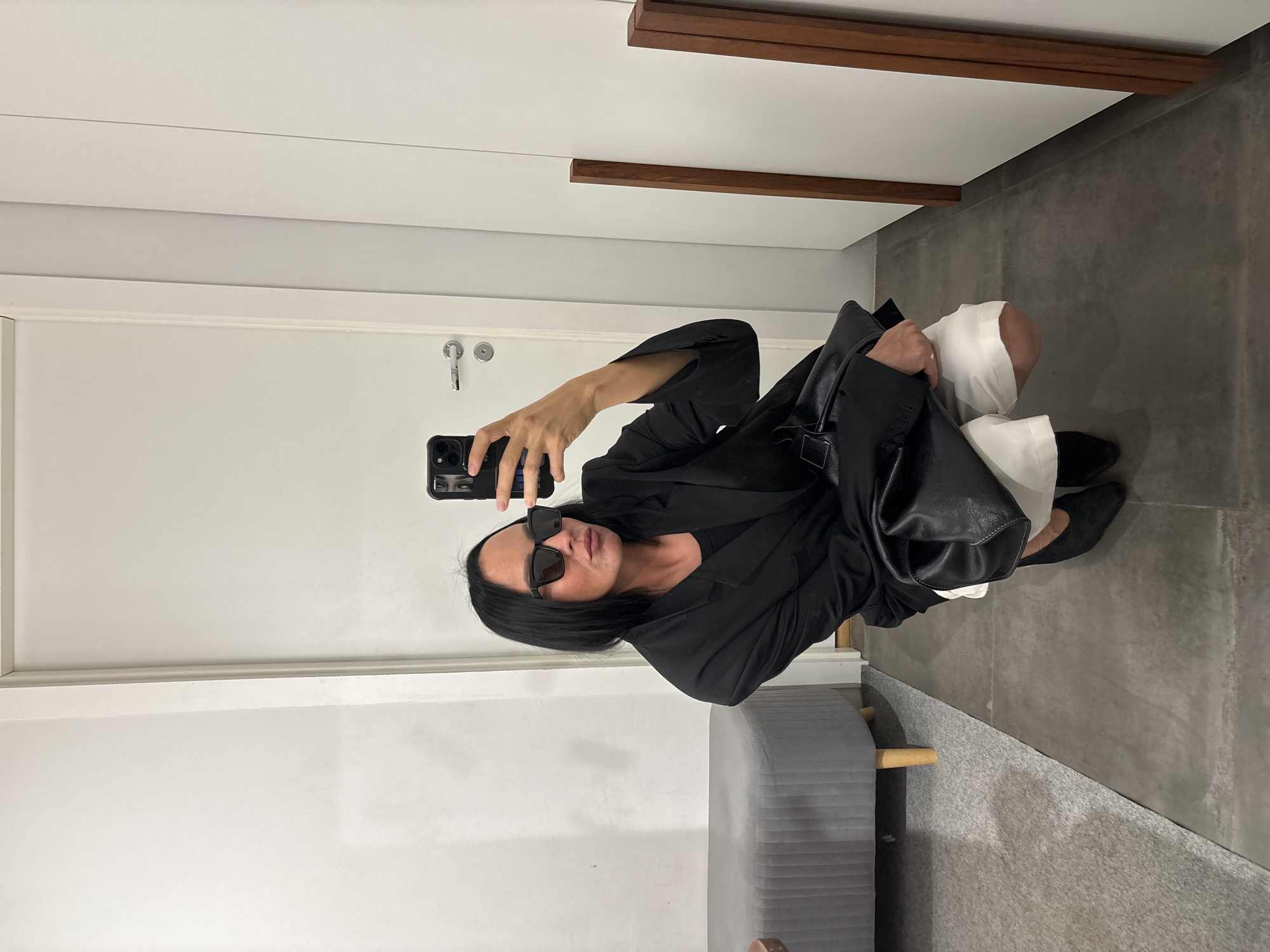 A woman is taking a selfie in a room with white walls and gray tiled flooring. She is wearing a black blazer, a black top, white shorts, black heeled shoes, and black sunglasses. She is holding a black smartphone in her right hand and a black handbag in her left arm. To her left, there is a grey upholstered bench with wooden legs. To her right, there are two wooden vertical beams. The woman is squatting and looking into the camera with her lips puckered. The location is not specified within the image and cannot be determined to be in Chaikovsky, Russia.