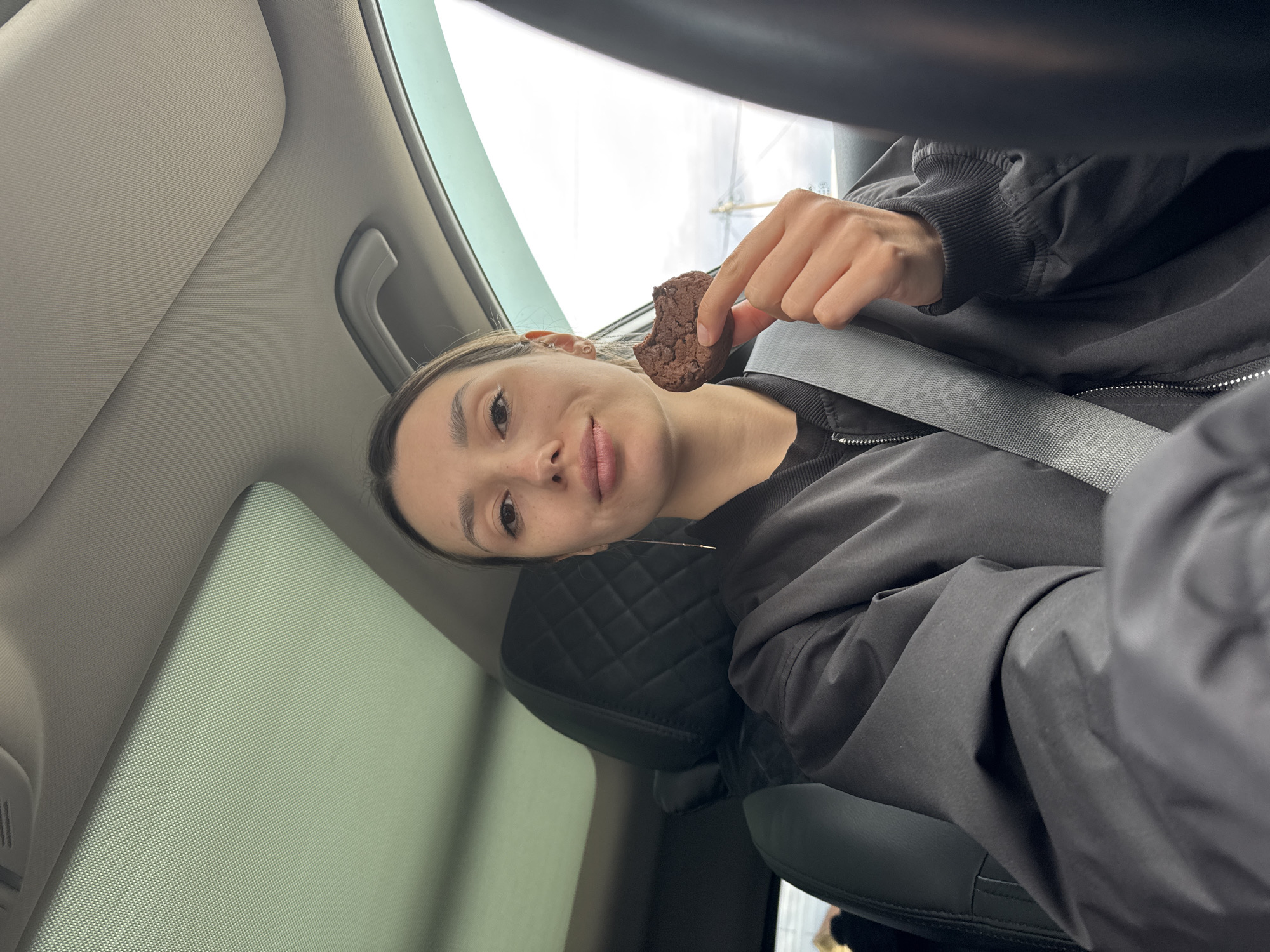 A young woman is depicted in a medium close-up shot, angled approximately 90 degrees clockwise from a standard upright orientation, suggesting the photo was taken from a tilted perspective or the camera was held sideways. She is seated inside a modern car, visible by the gray seatbelt across her chest and shoulder, the black quilted headrest behind her, and the beige/gray car interior elements like the headliner and sun visor.
The woman has fair skin, dark hair pulled back, and is wearing a dark, possibly black, jacket or sweatshirt over a dark top. She looks directly at the camera with a neutral to slightly smiling expression. Her left ear, which has multiple piercings, is visible.
She is holding a dark brown, possibly chocolate, cookie or brownie with a bite taken out of it, suggesting she is in the process of eating or has just taken a bite. Her right hand, holding the treat, is in the upper central part of the frame.
Through the car windows, a bright, diffuse light is visible, indicating it is daytime, possibly with an overcast sky. No specific outdoor scenery or landmarks are identifiable. There is no visible text in the image.