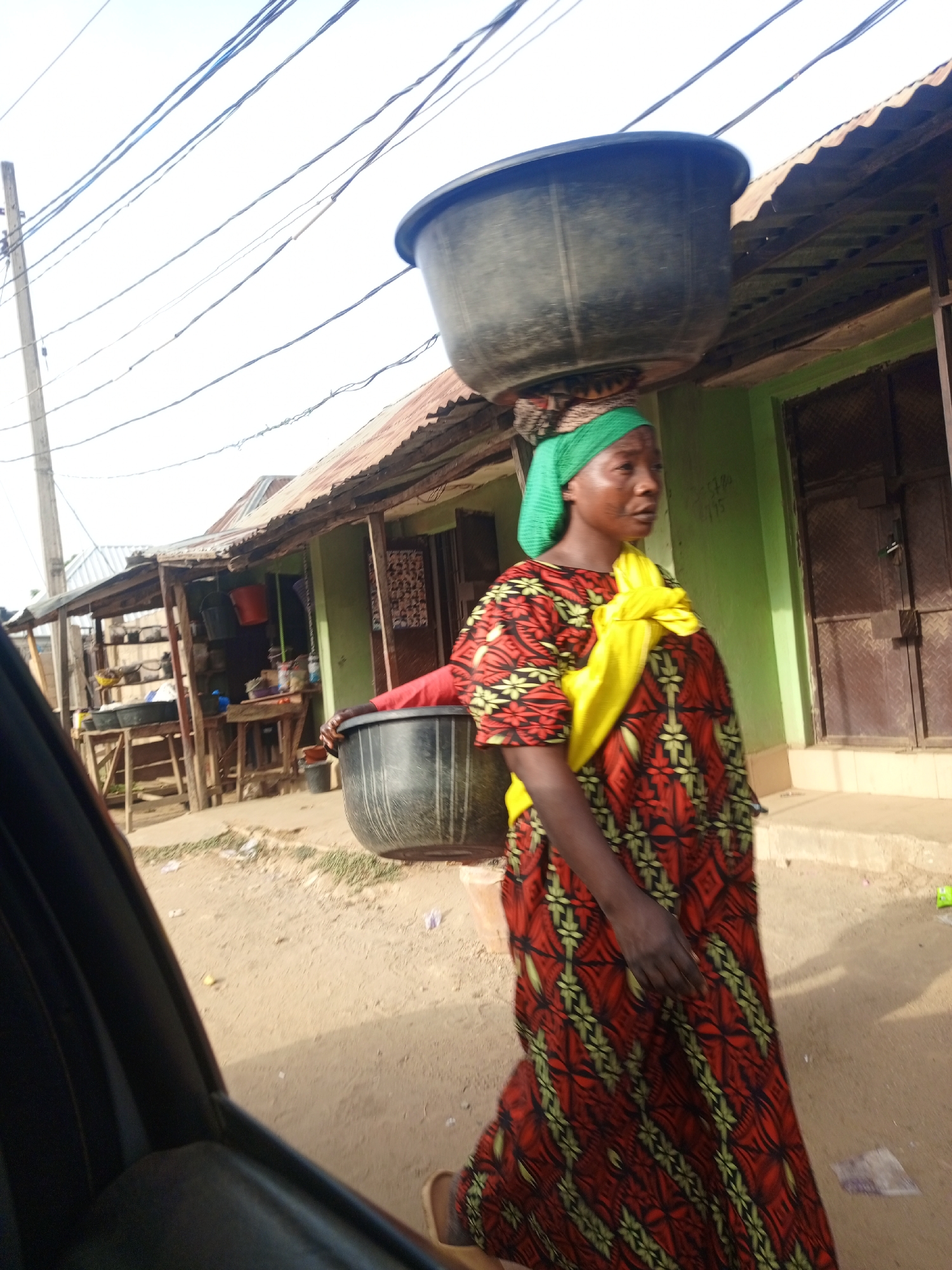 In an unidentified location in the city of Unknown, Unknown, a woman is walking along a dirt road. She is wearing a long, red and yellow patterned dress with a yellow cloth draped over her shoulder. She has a green headscarf on. On top of her head, she is carrying a large, black basin. In the background, there are buildings with corrugated metal roofs. A black basin sits outside of one building. There are utility poles and wires overhead. A portion of a dark vehicle is visible on the left side of the frame.