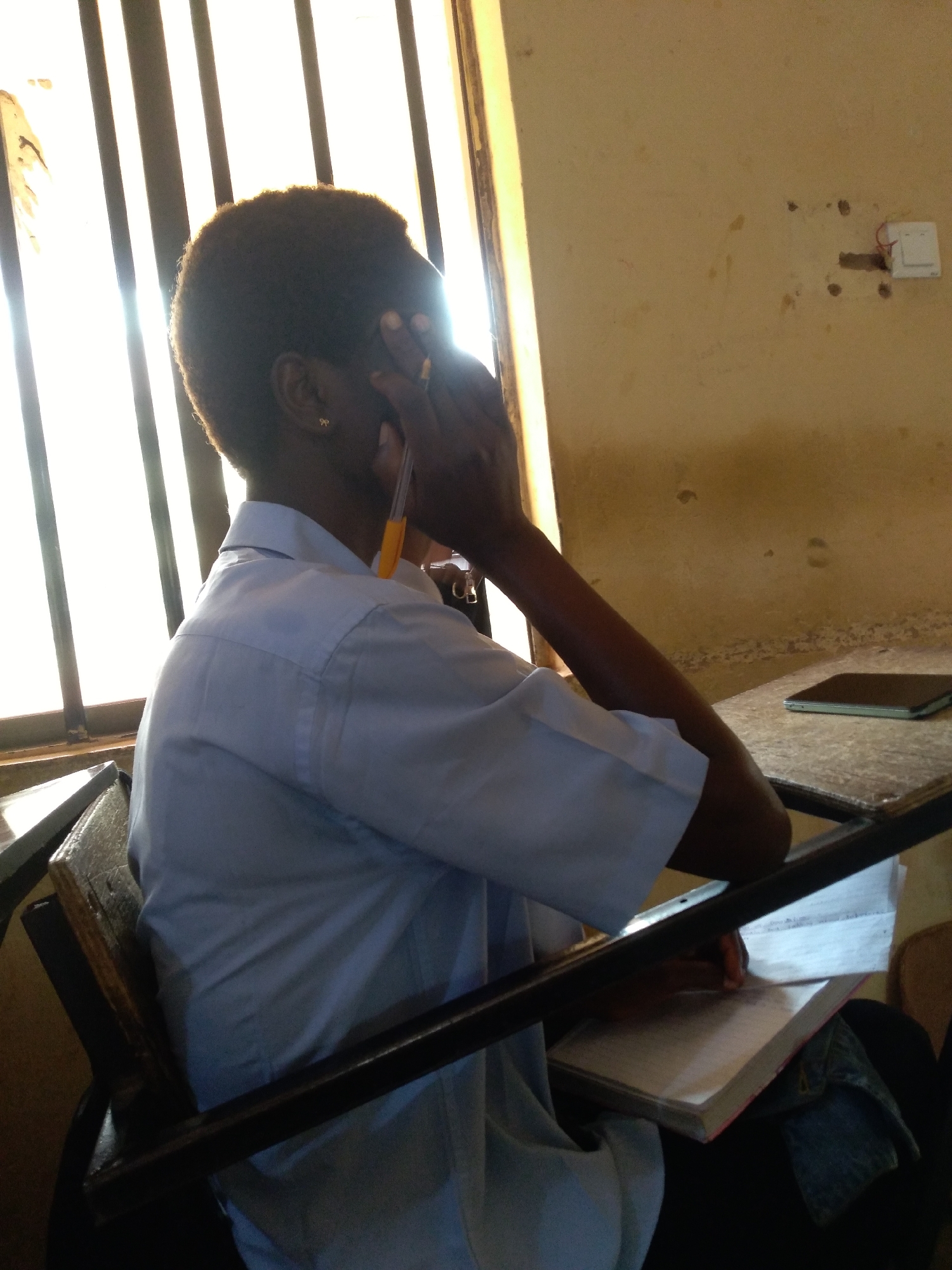 A young Black individual is seated at a desk, their body angled away from the camera, with their right hand covering their face. They are wearing a light blue collared shirt and have short, dark hair. A pen with an orange tip is visible between their fingers. In front of them on the desk is an open book with lined pages. To the right of the book, a dark rectangular object, possibly a tablet or e-reader, rests on the desk. The environment appears to be a classroom or study area with barred windows allowing bright sunlight to enter from the left. The walls are a muted yellowish-beige color, and a white electrical outlet with exposed red wires is visible on the right wall. The overall scene suggests a moment of concentration or fatigue during study. The presence of barred windows and the style of the electrical outlet could be indicative of educational institutions in Jos, Nigeria, where security measures like window bars are common, and electrical infrastructure can vary.
