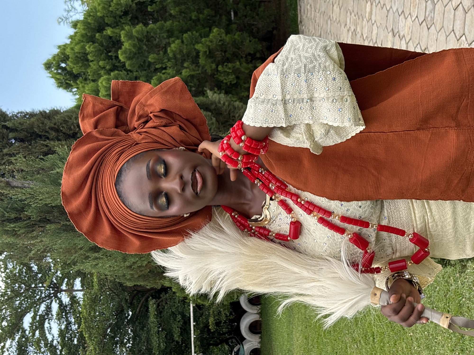 A Black woman, adorned in traditional attire and makeup, stands outdoors. She wears a vibrant orange headwrap, a cream-colored blouse with intricate white lace detailing and sequins, and a matching orange sash draped over her shoulder. She is accessorized with prominent red beaded necklaces and bracelets, and holds a white feathered fan in her left hand. The woman's eyes are closed, and her head is tilted slightly as she touches her face with her right hand. The background features lush green trees, a clear blue sky, and a paved walkway with hexagonal tiles. This scene is set in the city of Jos, Nigeria.