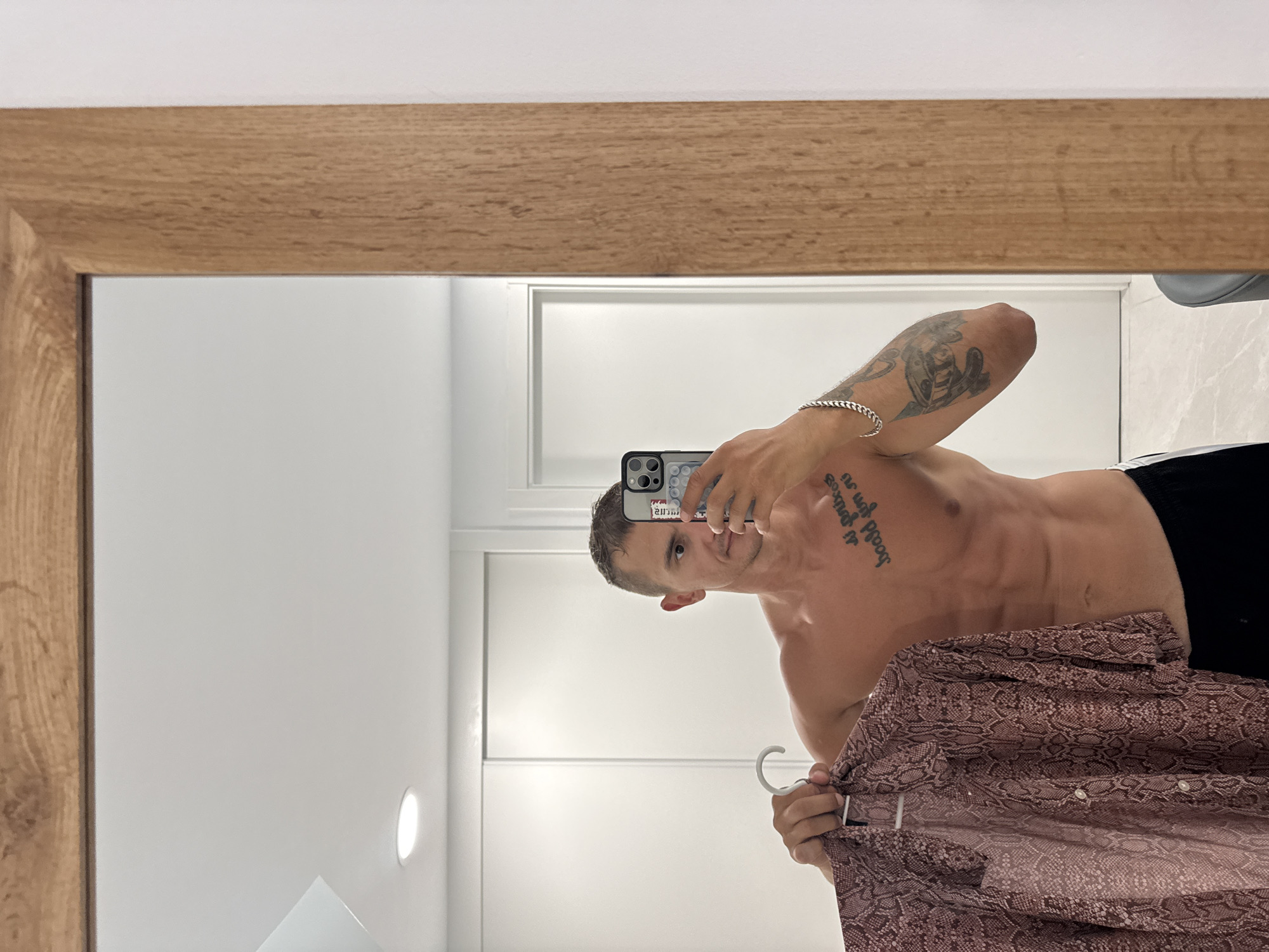 A shirtless man with visible abs and tattoos stands in front of a mirror, holding a snakeskin-patterned shirt on a hanger. He is in a brightly lit room with white sliding doors in the background. The mirror has a wooden frame. The scene appears to be a residential interior, possibly a bedroom or dressing area, located in Finestrat, Spain.