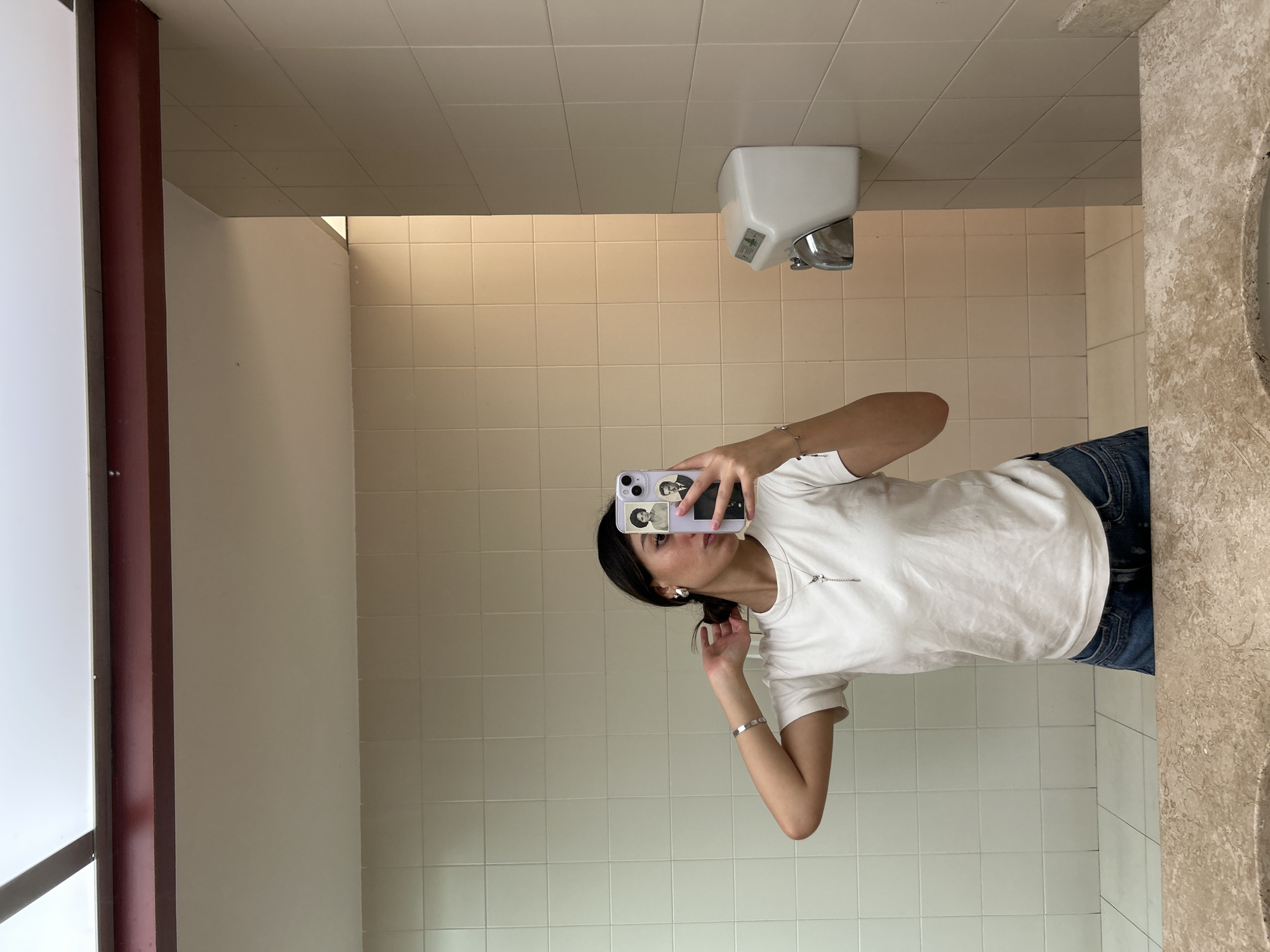 A person is seen in a mirror selfie, standing upright, in what appears to be a public restroom or similar tiled indoor space. The person, likely female, has dark hair pulled back and is wearing a plain white short-sleeved t-shirt, blue jeans, a silver bracelet on her left wrist, and a silver necklace with a small charm. She is holding a smartphone with a light purple case in her right hand, positioned to capture her reflection. The phone case has a few small, monochrome images or stickers on its back, one of which depicts a person's face.
The background of the mirror reflection shows a room with light beige or cream-colored square tiles covering the walls. The ceiling consists of light-colored acoustic panels. To the upper right of the person's reflection, a white, wall-mounted hand dryer with a chrome nozzle is visible. On the far right, a light-colored, mottled countertop or sink basin is partially reflected. A dark reddish-brown vertical beam or partition is seen on the far left of the image, contrasting with an adjacent white wall section. The lighting in the room is bright and even. The person's expression is neutral, focusing on taking the photograph. There is unreadable text or a logo on the hand dryer.