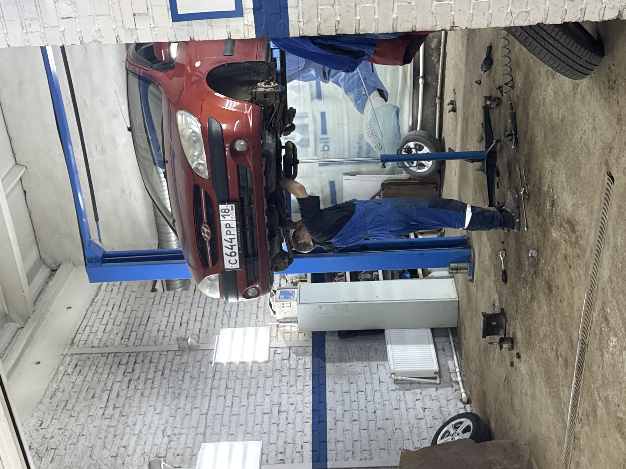 A mechanic wearing a black shirt and blue overalls is working on a red car lifted on a blue hydraulic lift in a garage. The car's front bumper displays a Russian license plate, "C644PP 18 RUSSIA." The garage has white brick walls, and various tools and car parts are scattered on the concrete floor. A tire leans against the wall in the background. Based on the license plate information, the scene is likely located in or around the city of Chaikovsky, Russia, as license plate region code 18 corresponds to the Udmurt Republic, where Chaikovsky is located.
