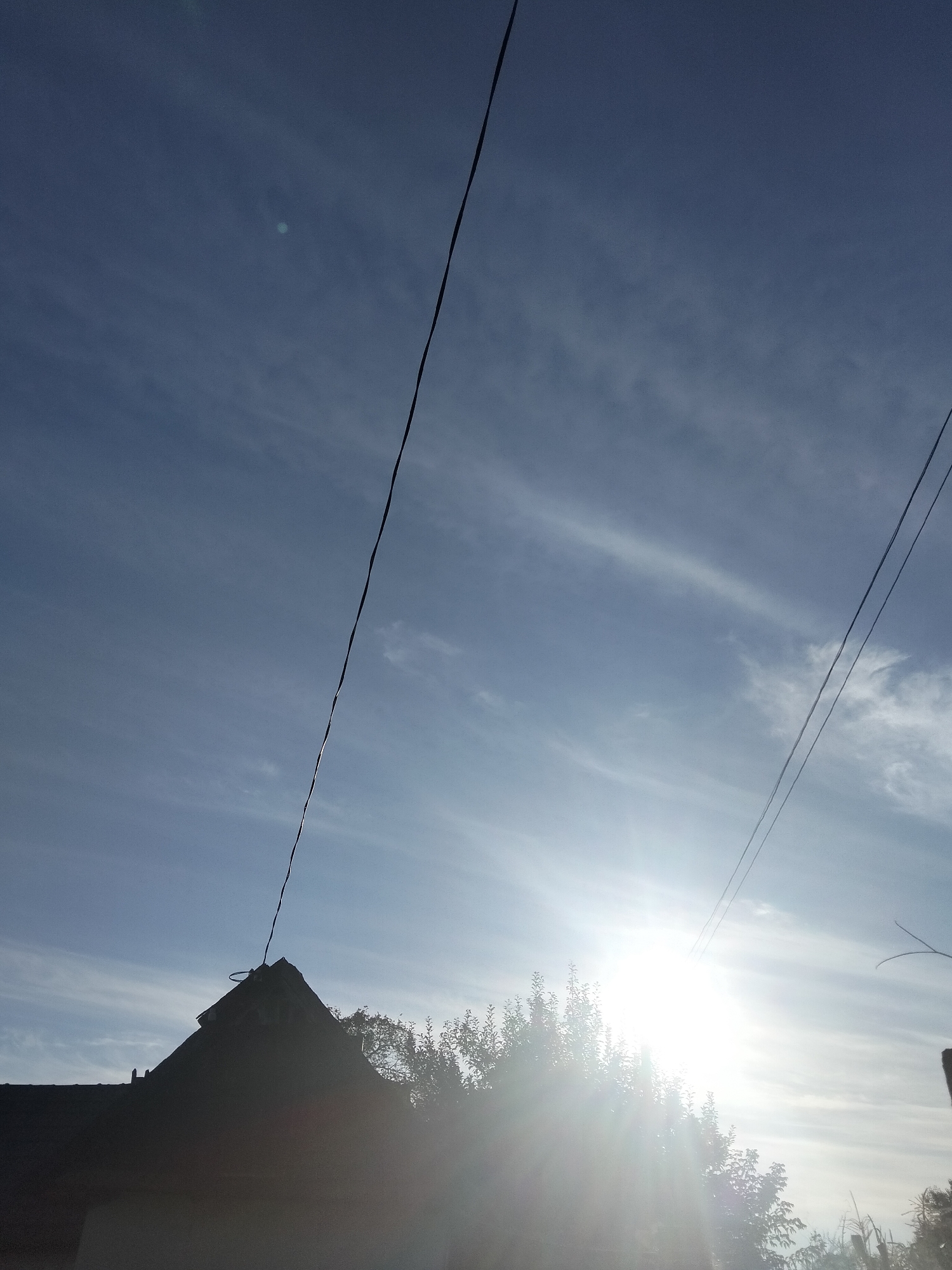 The image captures a low-angle view of a bright sun shining through a partly cloudy sky. Power lines crisscross the frame, silhouetted against the bright sky. In the foreground, the dark silhouette of a rooftop with a pointed gable is visible, partially obscured by lush green trees. The scene appears to be in the village of Kliushnykivka, Ukraine, suggesting a rural or suburban setting. The sunlight creates a lens flare, washing out some of the details in the upper right portion of the image.