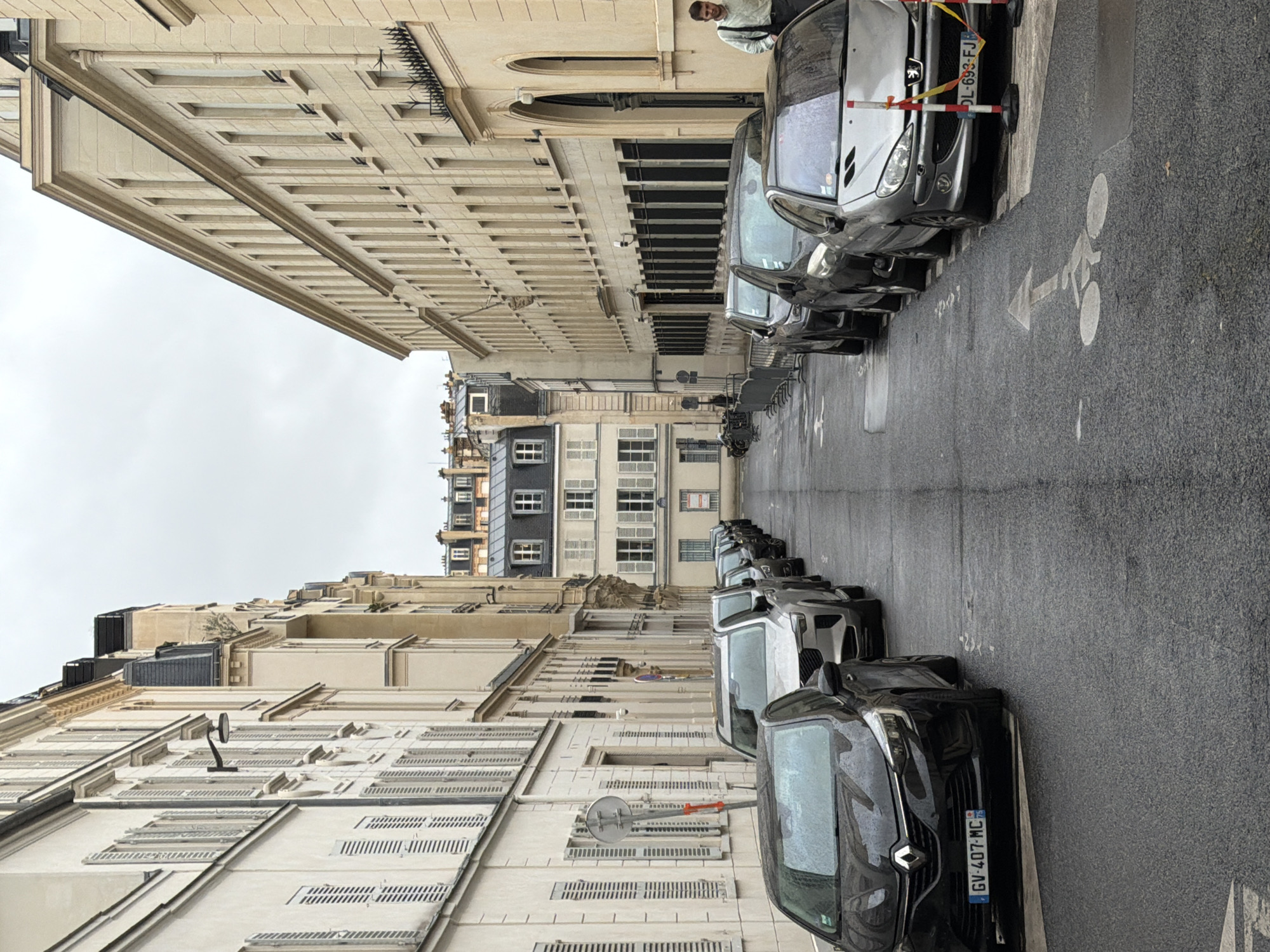 The image captures an upright view of a street scene in Paris, France, on what appears to be an overcast and possibly damp day, given the wet appearance of the asphalt road. Tall, classic Parisian buildings with light-colored stone facades, multiple stories, ornate cornices, and numerous windows line both sides of the relatively narrow street. Some buildings in the background feature distinctive mansard roofs.
The street is densely packed with parked cars along both sides. On the right side, a silver Peugeot is visible, bearing the French license plate "DL-693-FJ." Further down the street on the same side, a black Renault with the license plate "GV-407-MC" can be seen. Both license plates prominently display "75," indicating the Paris department. Red and white striped parking barriers are positioned in front of some parked vehicles.
On the road surface, a white bicycle symbol with an accompanying arrow is painted, indicating a dedicated or shared lane for cyclists. A single person, wearing a light-colored jacket or hoodie, stands on the sidewalk to the right, looking towards the camera. The architectural details of the buildings include decorative moldings, shuttered windows, and security features like bird spikes on ledges. The scene suggests a quiet, urban environment.