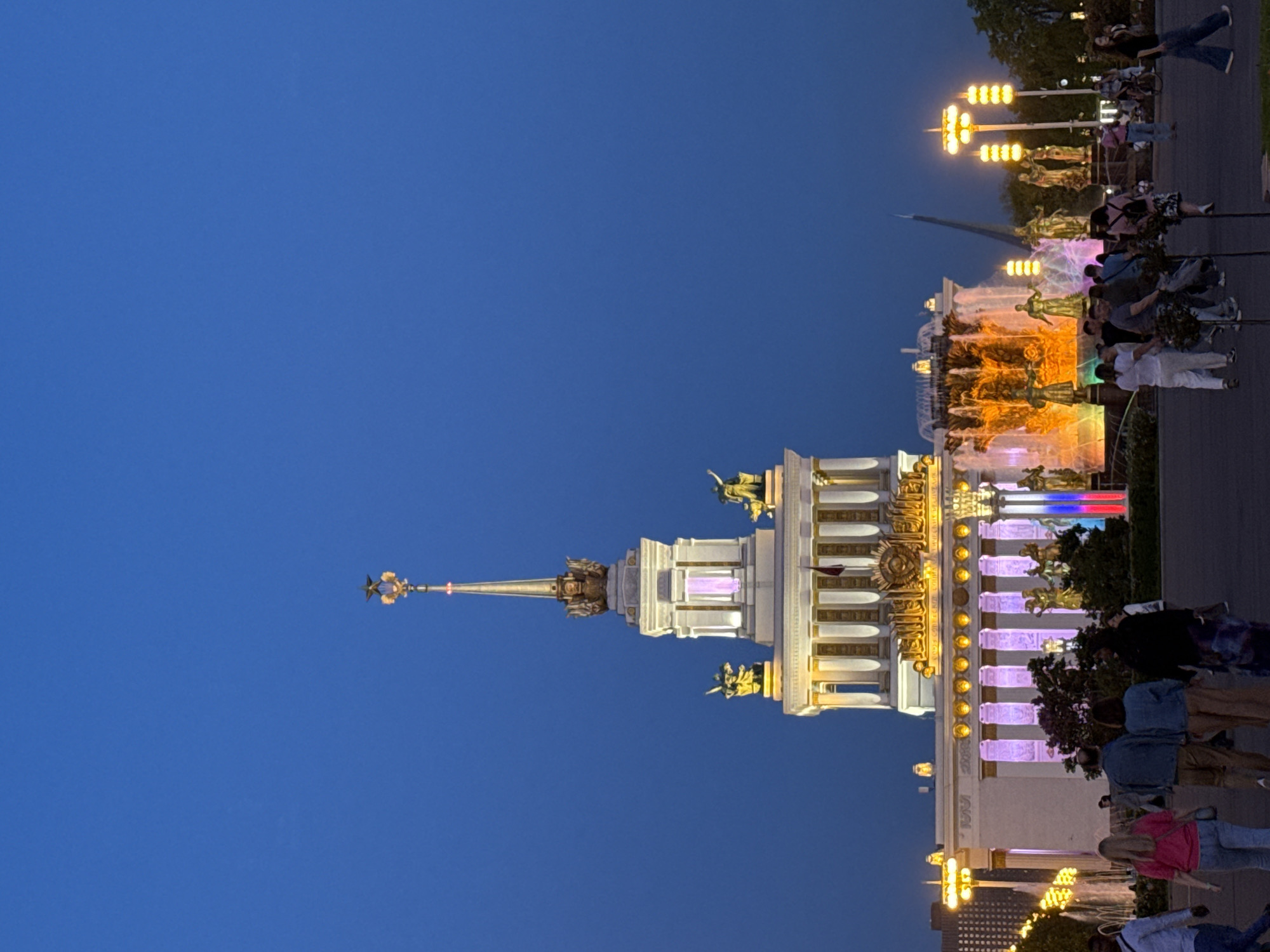 The image depicts a brightly lit building at night with a tall spire and statues, located in Moscow, Russia. The building is white and gold, with a Russian flag prominently displayed. People are walking near the building, and a fountain with colorful lights is visible. Streetlights are also illuminated. The sky is a deep blue.