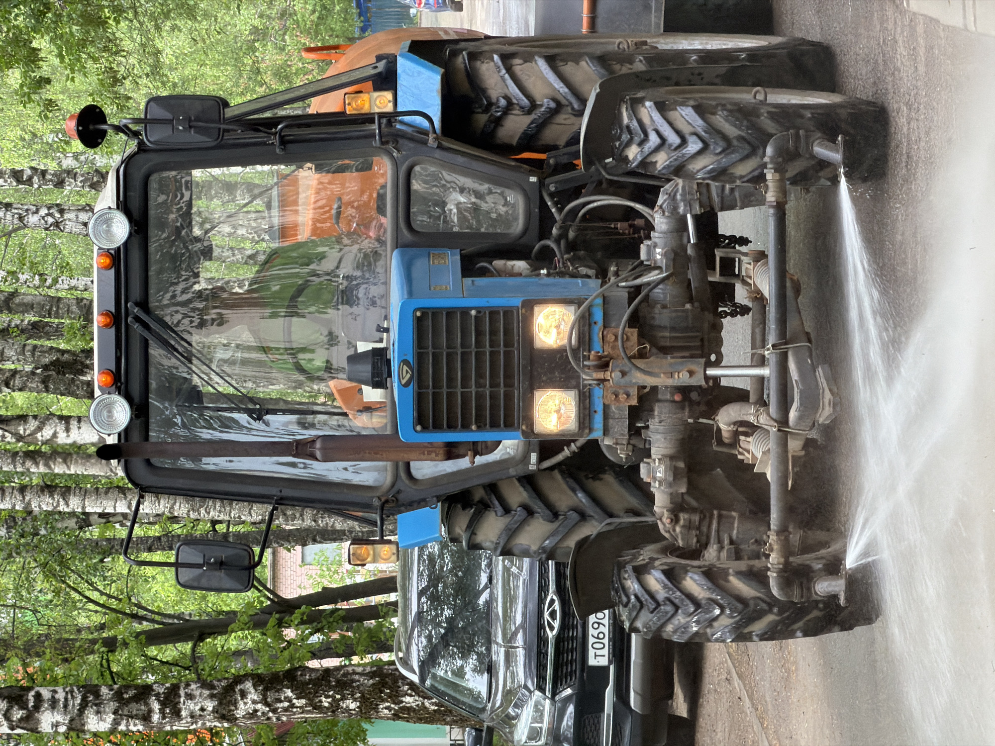 A tractor is parked on the edge of a paved area next to a Kia car, with water spraying out of a pipe attached to the front of the tractor. The tractor is a blue and orange MTZ tractor, with lights on and mud visible on the tires. The Kia is dark colored. Trees line the background. The license plate of the Kia is visible with the numbers T069OC. It’s likely a street scene in Dolgoprudny, Russia.