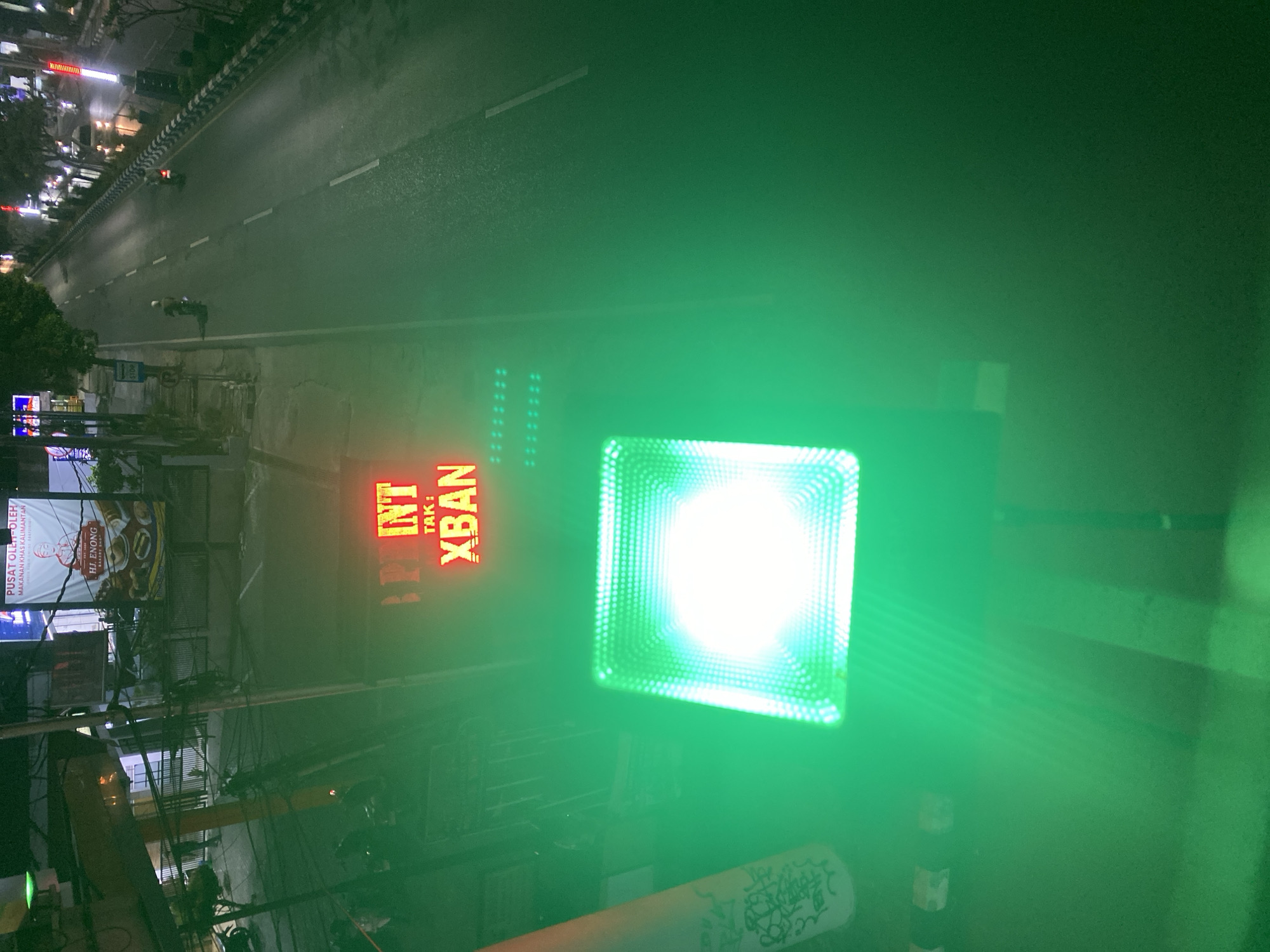 The image captures an outdoor nighttime scene in the city of Banjarbaru, Indonesia. In the foreground, a bright green LED light fixture is prominently displayed, emitting a strong glow. To the left of this fixture, a partially visible white cylindrical object with dark graffiti-like markings is present. Further back, a red LED sign displays the text "PINT TAK : XBAN". Above this sign, a banner with Indonesian text and an image of a person can be seen. The background features a street with several vehicles, including motorcycles, and streetlights illuminating the road. The environment appears to be an urban setting with overhead electrical wires visible, suggesting a developed area.
