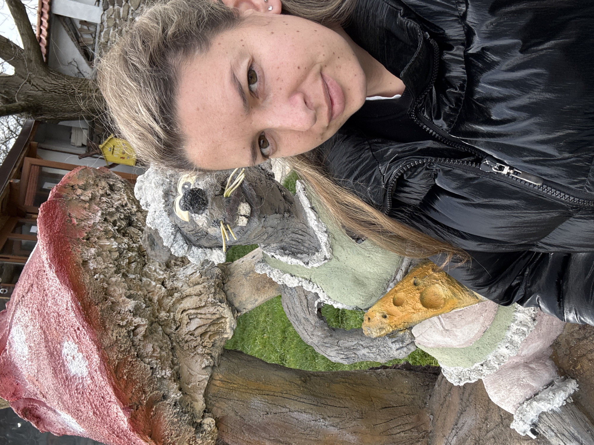 A woman in a black puffer jacket poses with a painted concrete sculpture of a mouse, situated outside a building. The sculpture includes a red and white mushroom-like structure and a piece of painted cheese. The image appears to be taken in Zlatitsa, Bulgaria, based on visual context clues. The woman's head is angled towards the camera, and she is smiling slightly. A house with a light-colored roof and bare branches of trees are visible in the background.