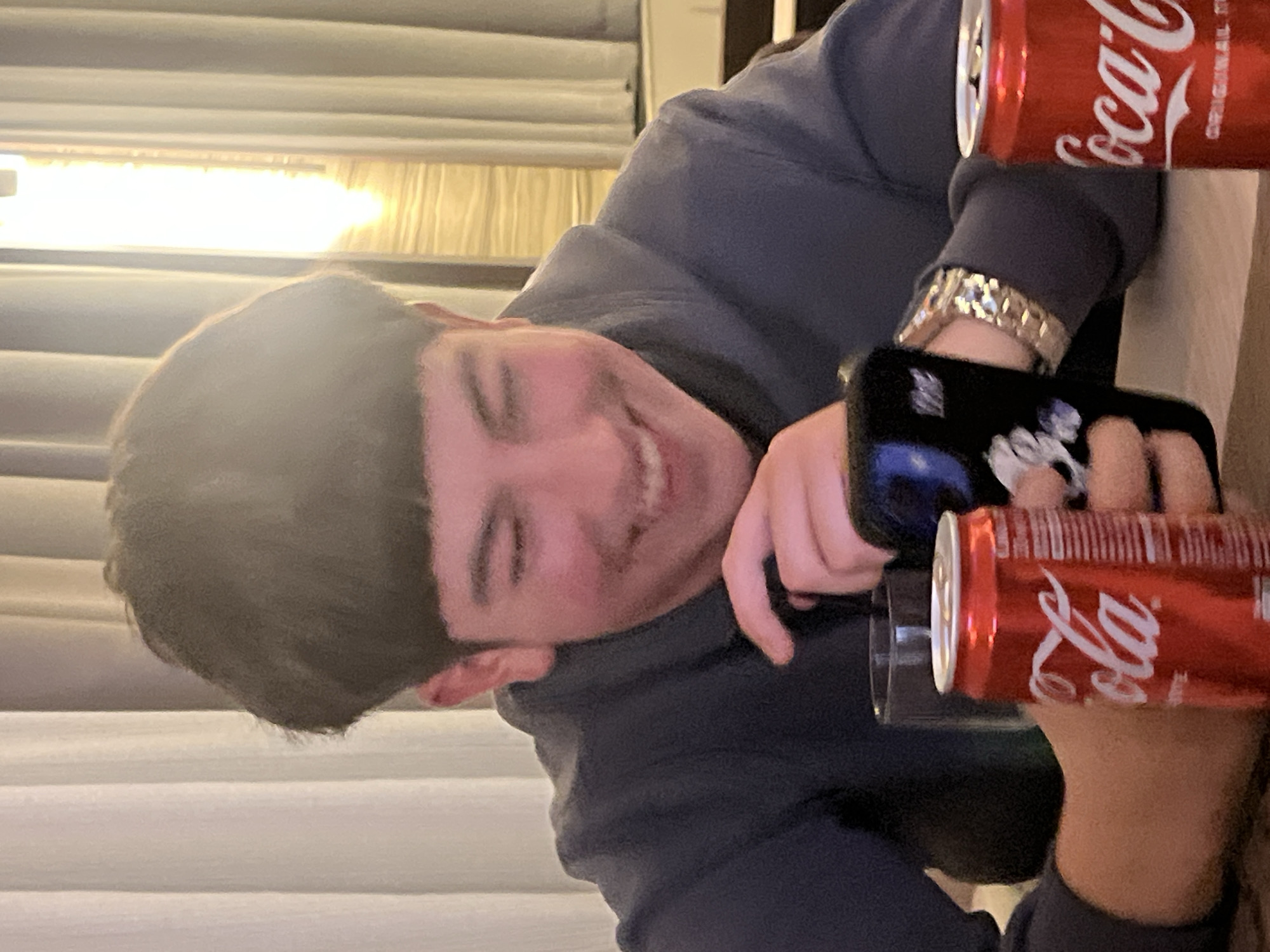 A photograph taken from a slightly upward angle captures a young person, likely male, smiling broadly at the camera. They have short, dark hair and are wearing a dark blue or navy hoodie/sweatshirt.
The individual is holding a dark-colored smartphone with both hands, its screen or case showing some white graphic elements. On their left wrist, a gold-colored, chunky watch or bracelet is visible. Two red Coca-Cola cans are prominent in the image; one is partially visible in the upper right, and another is held lower down, near the phone, with a clear glass beneath it. The cans clearly display the "Coca-Cola" logo and "ORIGINAL" text.
The setting appears to be indoors. In the background on the left, bright, warm light streams through vertical blinds, suggesting either a window during daylight or a strong artificial light source. The right side of the background is less distinct, possibly a wall or part of a furniture piece.
The person's wide smile conveys a sense of happiness or amusement, indicating a relaxed and informal moment. The activity suggests a casual social interaction or a moment of leisure. The image is oriented sideways, with the person's head rotated 90 degrees to the left, making them appear to be looking upwards in the current frame. The provided location context for this scene is Kochan, Bulgaria.
