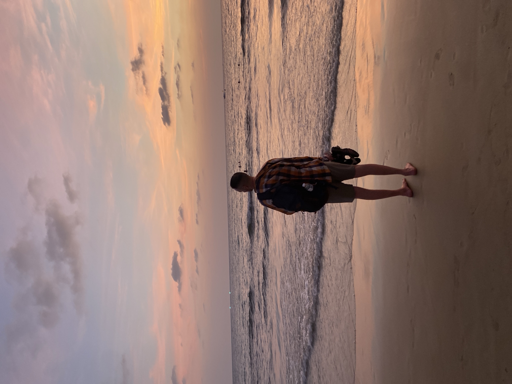The image shows a man standing on a beach at dusk. He is wearing a plaid shirt, shorts, and a backpack, and appears to be barefoot. The tide is coming in, and small waves are visible in the water. The sky is a mix of pink, orange, and gray clouds. The photo was taken at Karon Beach, Thailand.