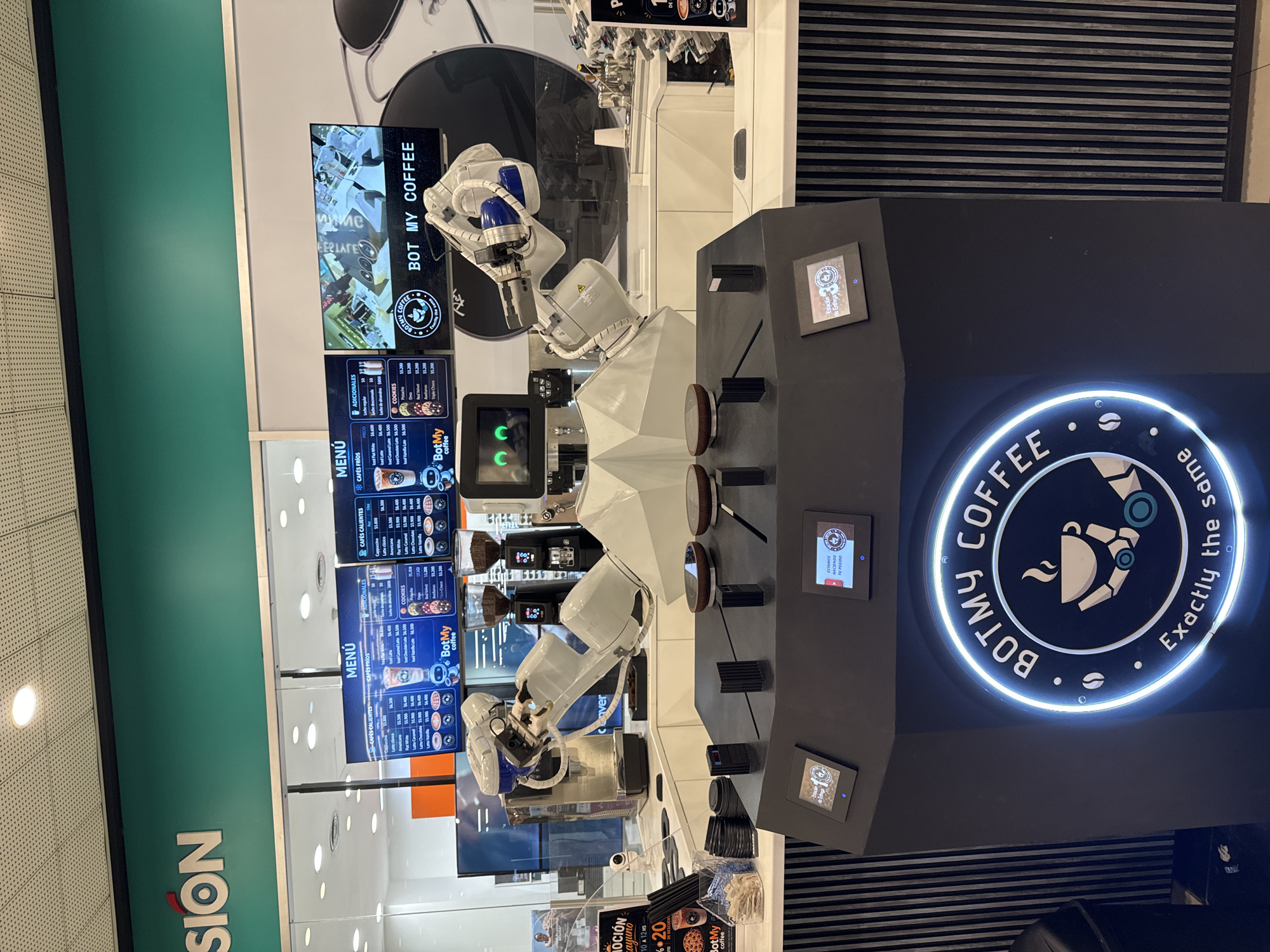 The image shows a BotMy Coffee automated coffee station. The station features robotic arms that prepare coffee, menu screens displaying options and prices, and a prominent illuminated logo. A green wall and visible signage suggest the station is located in a retail or commercial environment. While the location isn't explicitly stated, the presence of Spanish-language signage and currency symbols (e.g., "$") indicates it could be in Buenos Aires, Argentina.