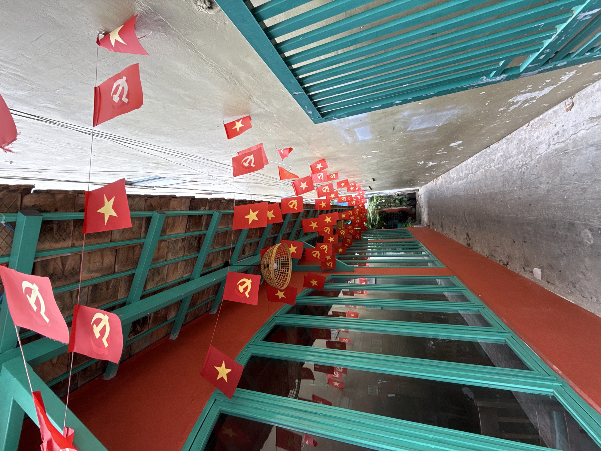 This image depicts a narrow alleyway in Nha Trang, Vietnam, lined with buildings adorned with red flags featuring yellow stars and hammers and sickles, symbols of communism. The flags are strung along lines, creating a festive or celebratory atmosphere. On the left, a building with bright orange walls features large, turquoise-framed windows. The alley floor is paved with concrete. On the right, a weathered, pale yellow wall stands next to a vibrant teal metal fence. The scene suggests a cultural or historical context, possibly related to a national holiday or political commemoration in Vietnam.